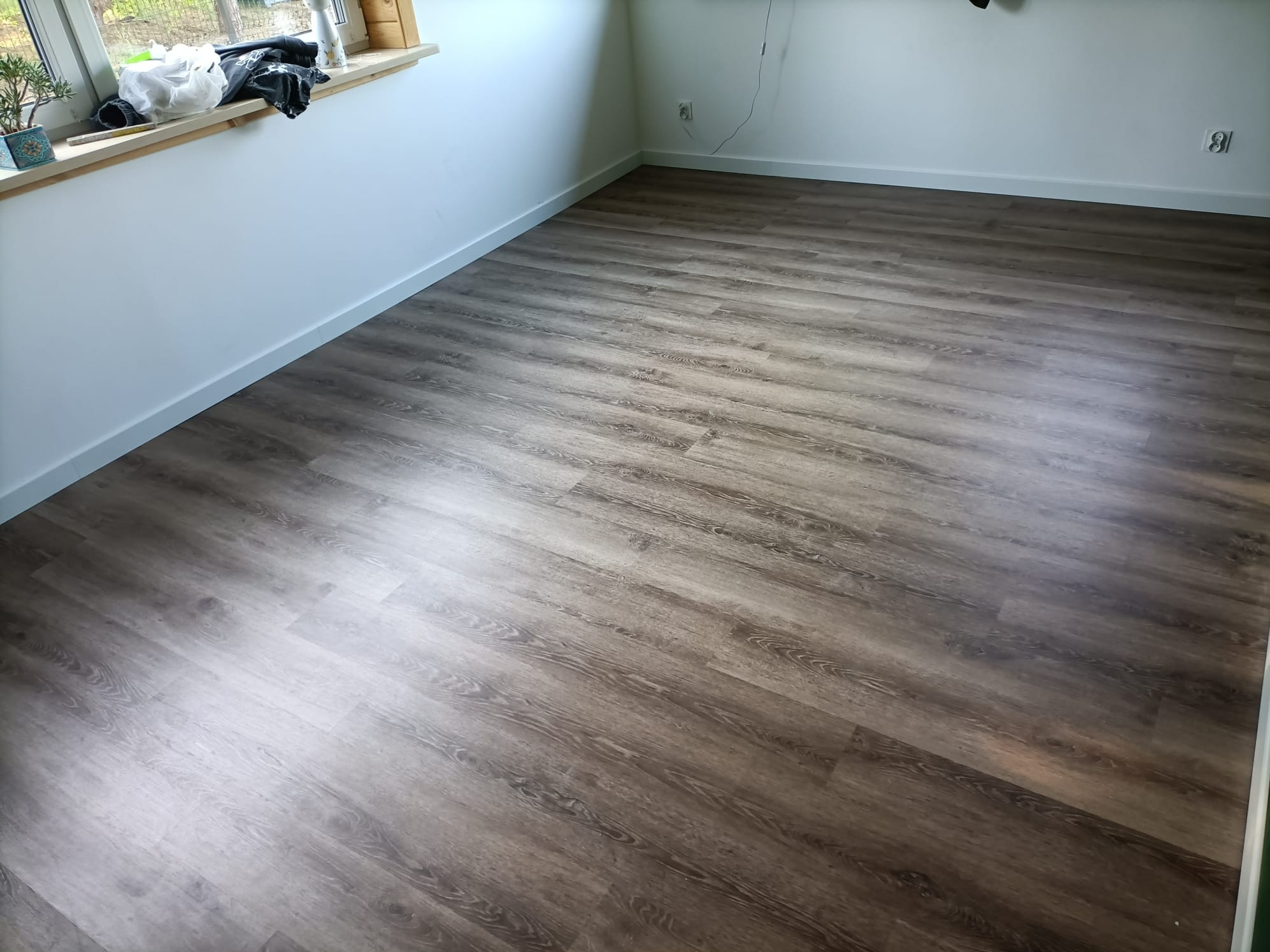 Ukończona podłoga z paneli Fatra Marillo w jasnym pomieszczeniu, widok z góry, listwy przypodłogowe w kolorze ścian, na parapecie doniczka z rośliną i przedmioty.