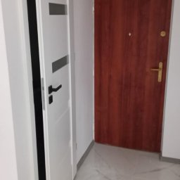 Paweł Szerszeń - Korytarz po remoncie z dwiema drzwiami: białe drzwi wewnętrzne z czarną klamką i brązowe drzwi wejściowe z złotą klamką. Białe ściany i marmurowa podłoga.