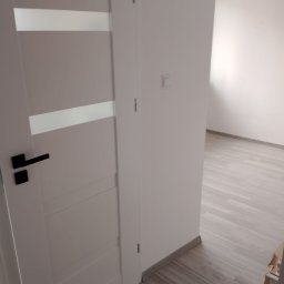 Paweł Szerszeń - Białe drzwi wewnętrzne z czarną klamką i poziomymi przeszkleniami, widok na wykończone wnętrze z panelami podłogowymi.