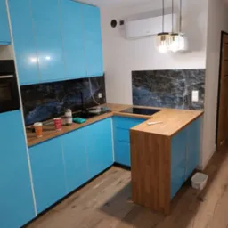 Niebieska, lakierowana zabudowa kuchenna z drewnianym blatem, czarnym panelem nad blatem i nowoczesnym oświetleniem wiszącym, widok na fragment drewnianej podłogi.