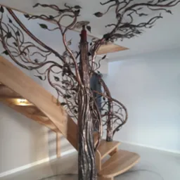 Kuta balustrada schodowa w kształcie drzewa z metalowymi liśćmi, łącząca się z drewnianymi schodami w jasnym wnętrzu.