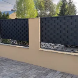 Nowoczesne ogrodzenie z czarnych, plecionych paneli metalowych i dekoracyjnymi, kutymi elementami w kształcie liści, osadzone na niskim, beżowym murku z kamiennymi słupkami po lewej stronie.