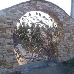 ADK blacksmith CNC Adam Kłos - Kuta brama w kształcie drzewa z liśćmi, osadzona w kamiennym łuku z jasnych, nieregularnych kamieni.