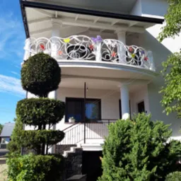 Biała, ażurowa balustrada balkonowa z kolorowymi motylami i kwiatami na tle białego domu z kolumnami i zadbaną zielenią.