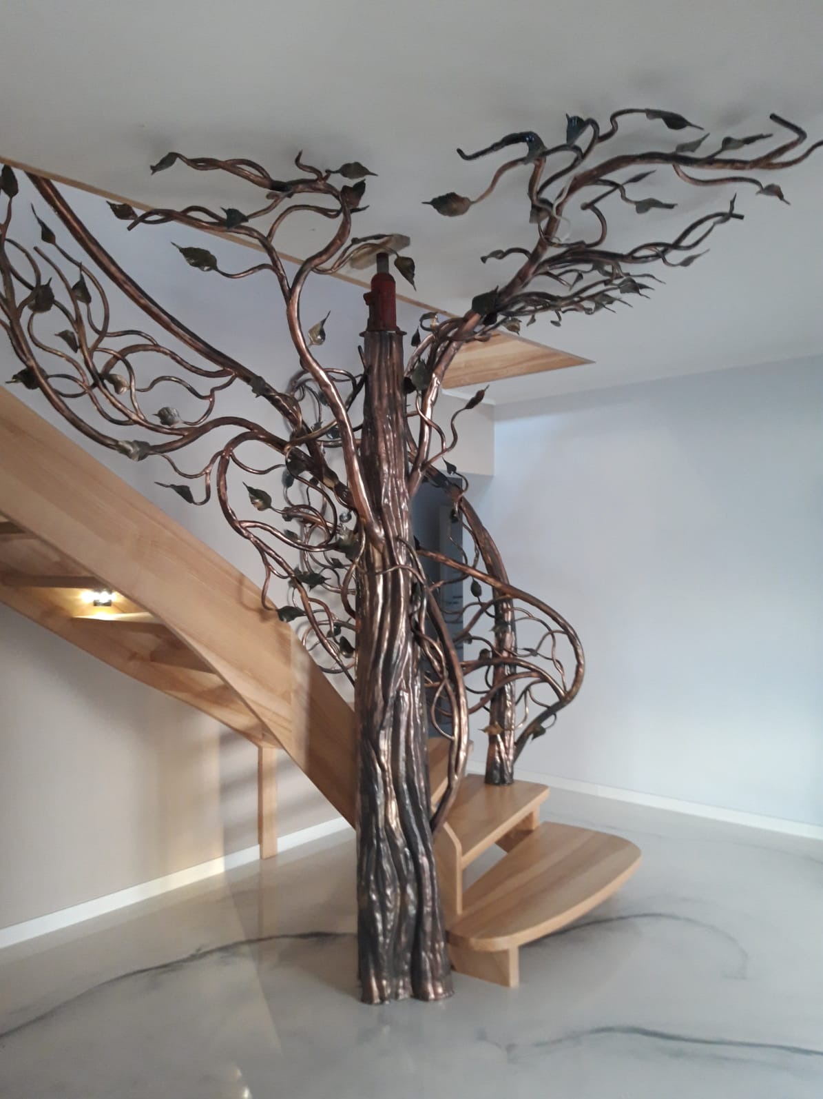 Kuta balustrada schodowa w kształcie drzewa z metalowymi liśćmi, łącząca się z drewnianymi schodami w jasnym wnętrzu.