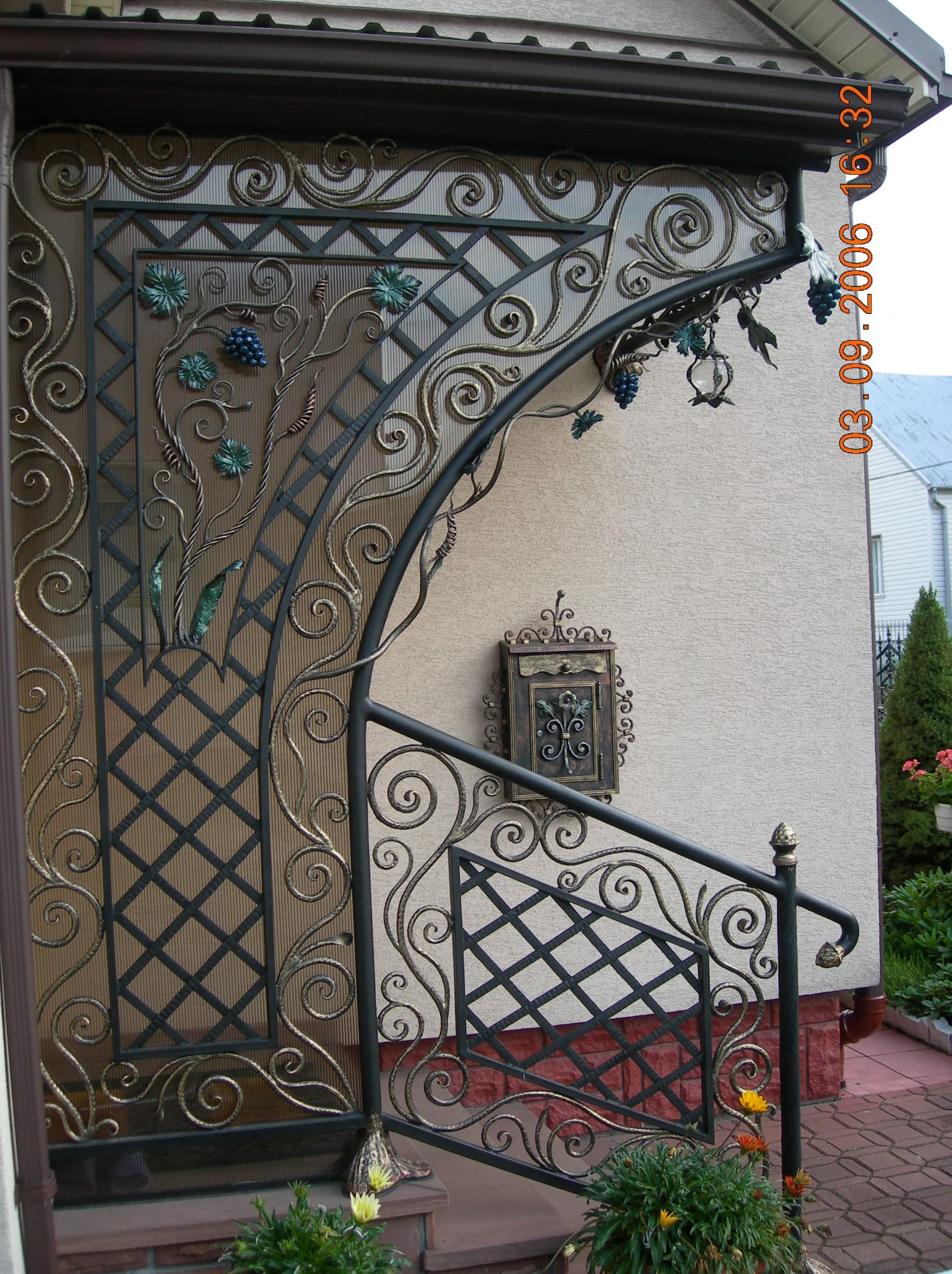 Kute zadaszenie nad wejściem z motywem winogron, zdobiona skrzynka na listy i fragment balustrady z kutymi elementami na tle elewacji domu.