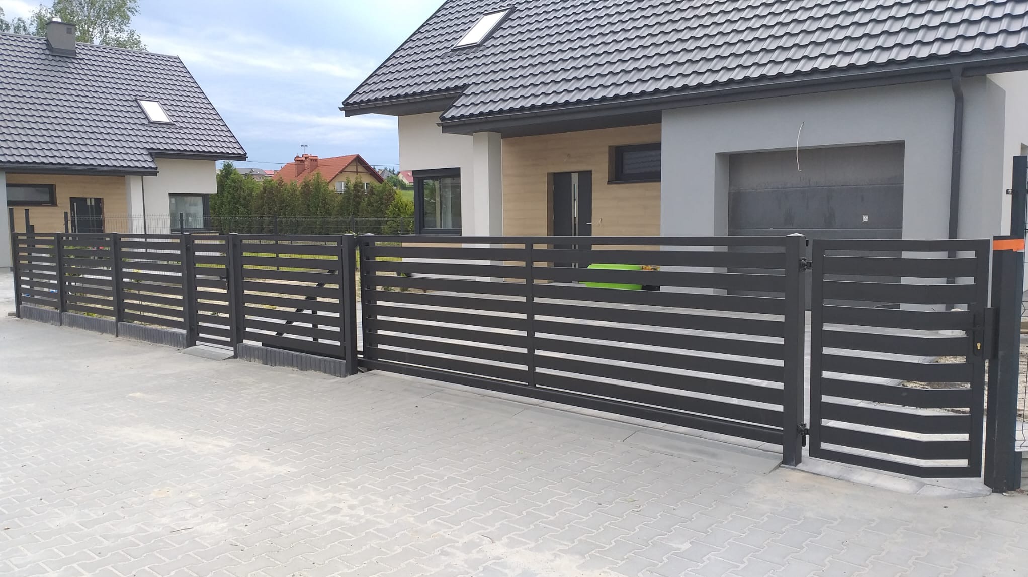 Nowoczesne, poziome ogrodzenie panelowe w kolorze antracytowym, z furtką i bramą wjazdową, na tle domu z szarą elewacją i grafitowym dachem.