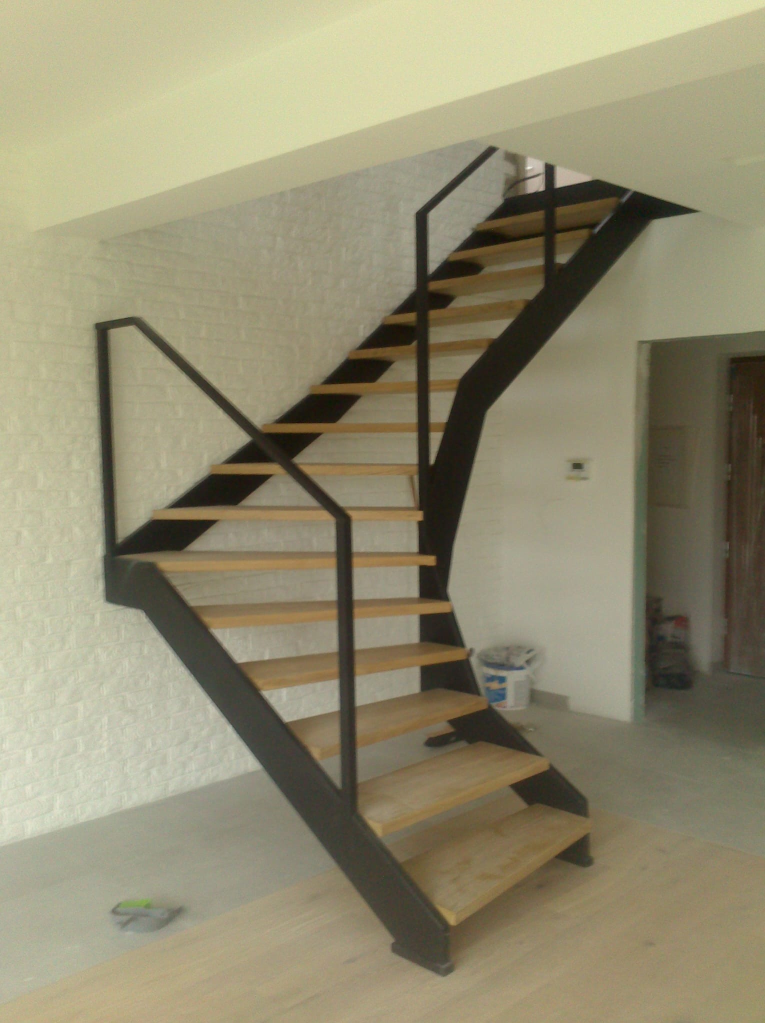 Metalowe schody z drewnianymi stopniami i czarną balustradą, zamontowane w jasnym wnętrzu z białą ceglaną ścianą. W tle widoczne wiadro i otwarte drzwi.