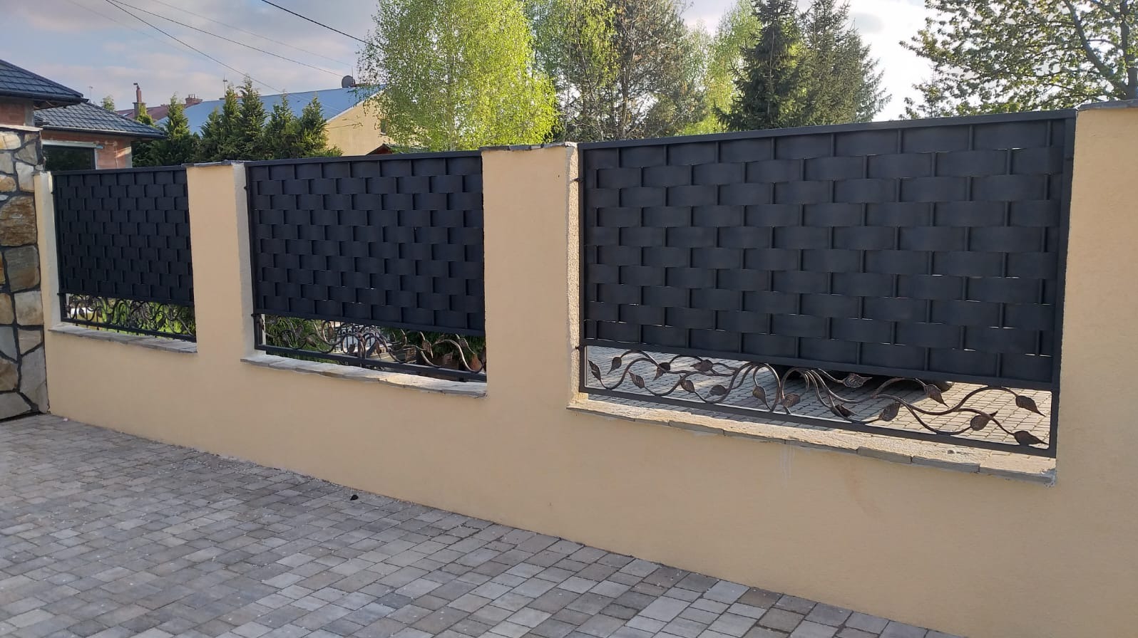 Nowoczesne ogrodzenie z czarnych, plecionych paneli metalowych i dekoracyjnymi, kutymi elementami w kształcie liści, osadzone na niskim, beżowym murku z kamiennymi słupkami po lewej stronie.