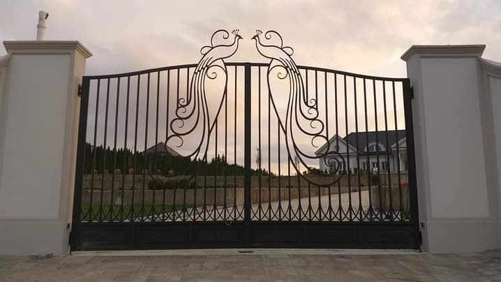 Kuta brama wjazdowa z motywem dwóch pawi, osadzona na masywnych, jasnych słupach, z widokiem na dom w tle.