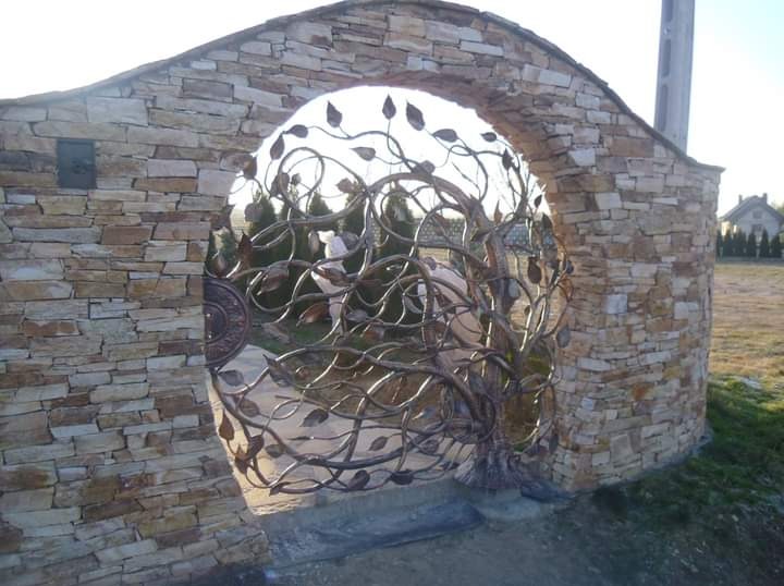 Kuta brama w kształcie drzewa z liśćmi, osadzona w kamiennym łuku z jasnych, nieregularnych kamieni.