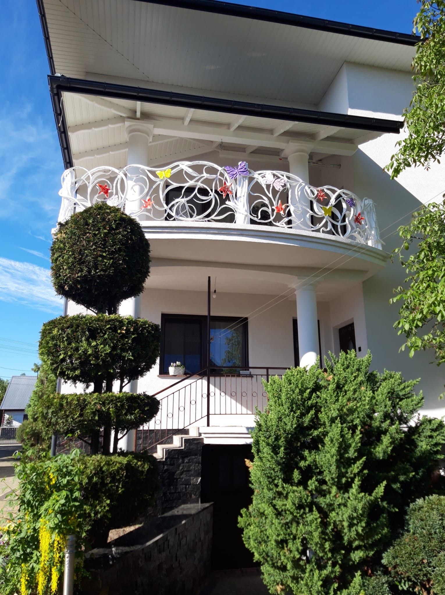 Biała, ażurowa balustrada balkonowa z kolorowymi motylami i kwiatami na tle białego domu z kolumnami i zadbaną zielenią.