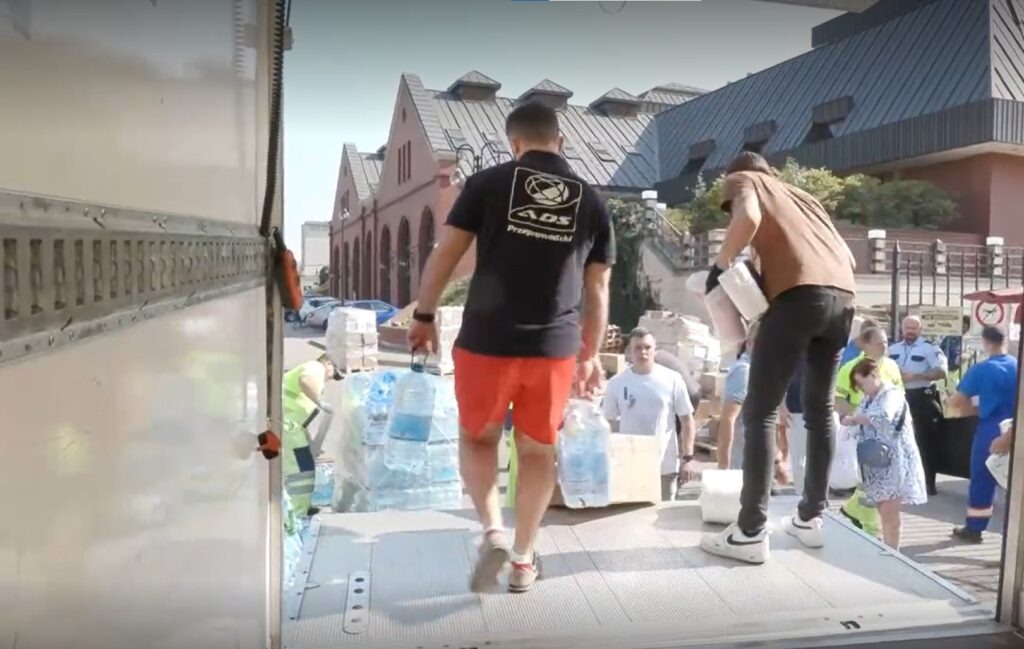 Rozładunek butelkowanej wody z ciężarówki, widoczni pracownicy w odblaskowych kamizelkach i osoby cywilne, w tle budynek z czerwonej cegły.