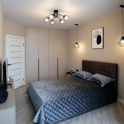 SN MEBLE SPÓŁKA Z OGRANICZONĄ ODPOWIEDZIALNOŚCIĄ - Nowoczesna sypialnia z zabudowaną szafą o gładkich, beżowych frontach, łóżkiem z pikowaną narzutą w odcieniach szarości i brązowym zagłówkiem, oświetlona wiszącymi lampami w stylu industrialnym...