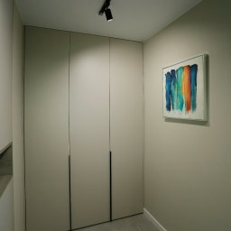 SN MEBLE SPÓŁKA Z OGRANICZONĄ ODPOWIEDZIALNOŚCIĄ - Beżowa zabudowa szafy z minimalistycznymi, czarnymi uchwytami, oświetlona reflektorem szynowym; na ścianie abstrakcyjny obraz z pionowymi smugami w odcieniach niebieskiego, zielonego i pomarańczowego.