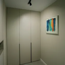 Beżowa zabudowa szafy z minimalistycznymi, czarnymi uchwytami, oświetlona reflektorem szynowym; na ścianie abstrakcyjny obraz z pionowymi smugami w odcieniach niebieskiego, zielonego i pomarańczowego.