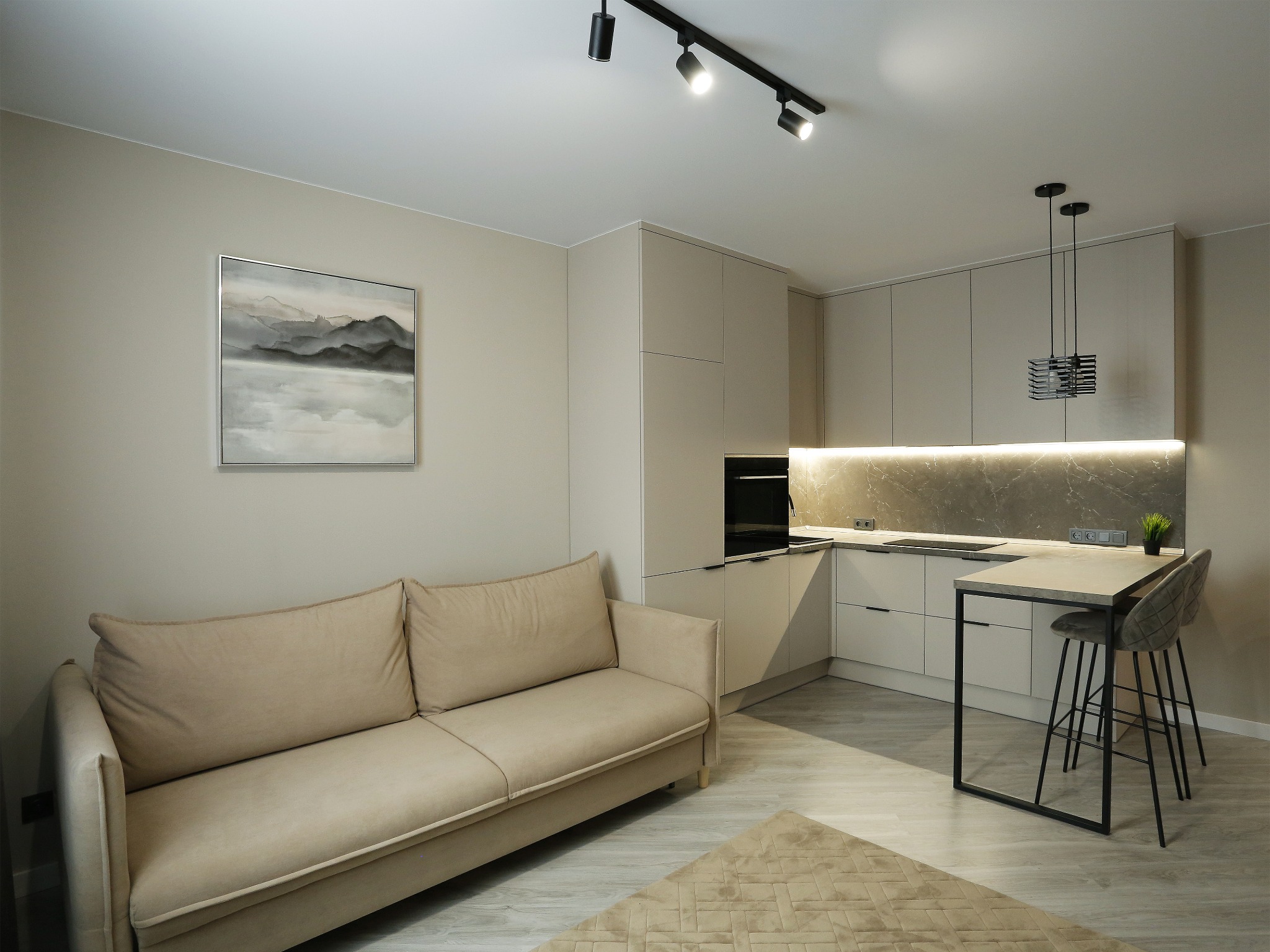 Jasne, minimalistyczne wnętrze z beżową sofą, obrazem górskim na ścianie i aneksem kuchennym z wyspą i hokerami, oświetlone reflektorami i lampami wiszącymi.