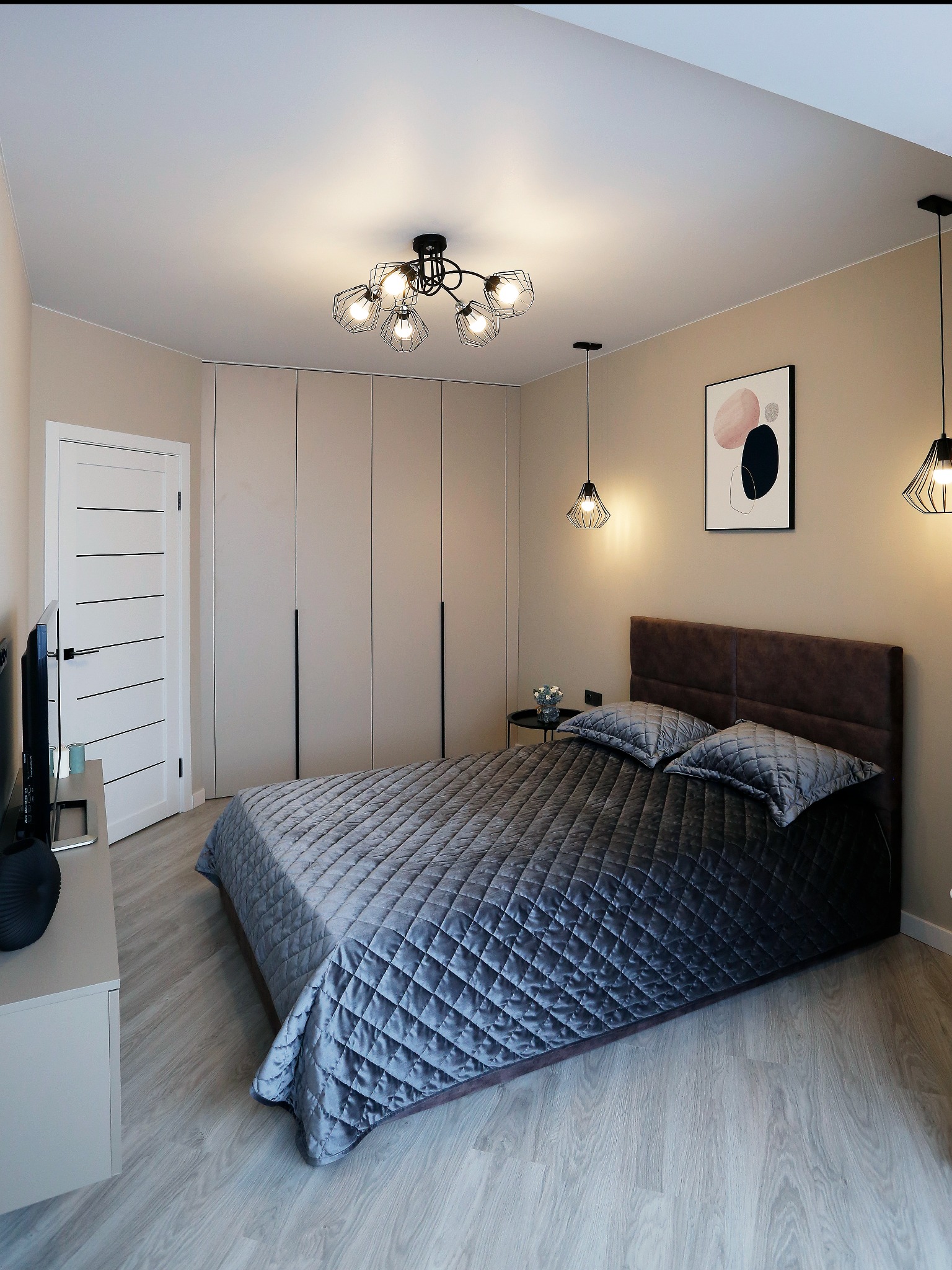 Nowoczesna sypialnia z zabudowaną szafą o gładkich, beżowych frontach, łóżkiem z pikowaną narzutą w odcieniach szarości i brązowym zagłówkiem, oświetlona wiszącymi lampami w stylu industrialnym...