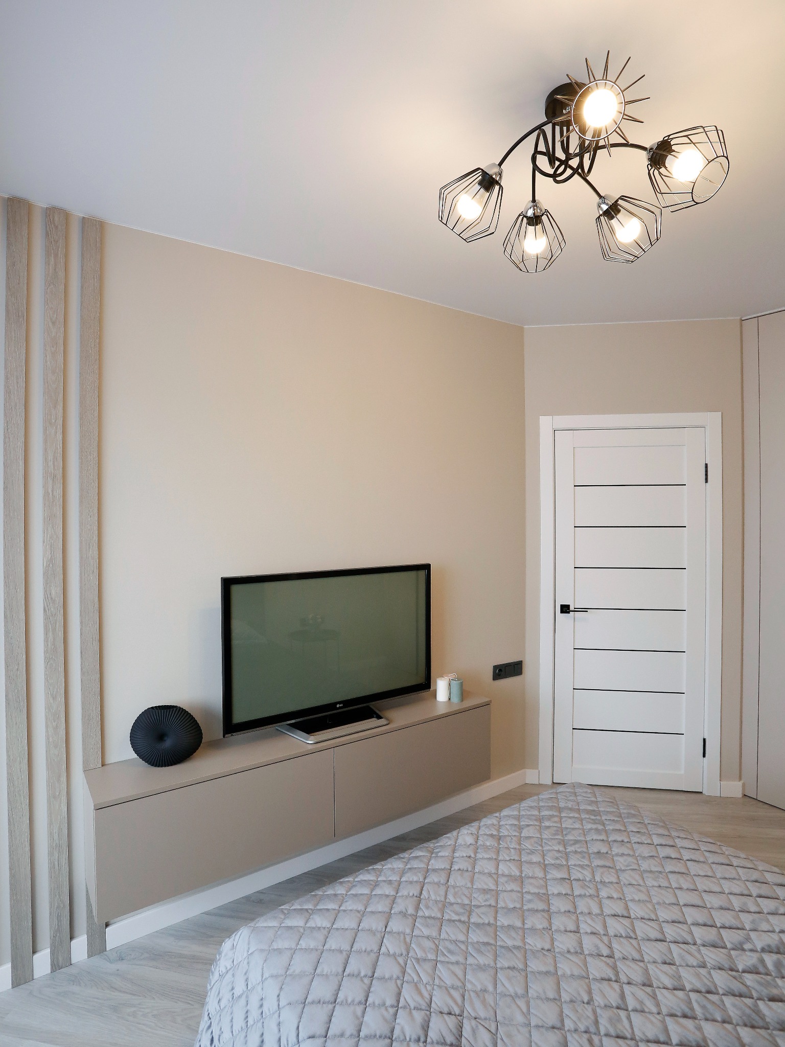 Nowoczesna sypialnia w beżowych tonacjach z podwieszaną szafką RTV, telewizorem, dekoracyjnym panelem ściennym z drewnianymi lamelami, białą, minimalistyczną drzwią i designerską lampą sufitową.
