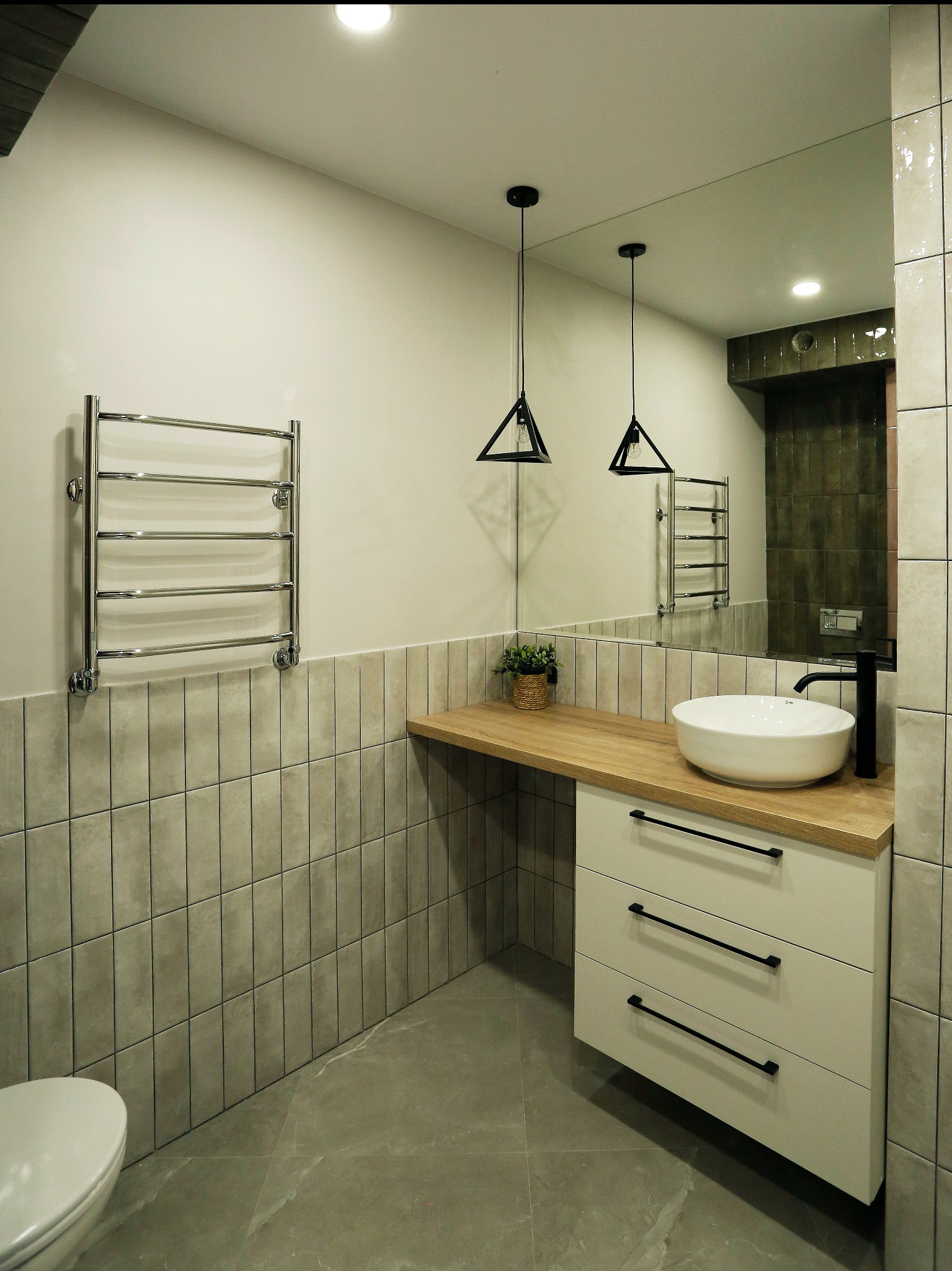 Nowoczesna łazienka z umywalką nablatową, szafką z trzema szufladami z czarnymi uchwytami, drewnianym blatem, grzejnikiem drabinkowym i wiszącymi lampami o geometrycznych kloszach.