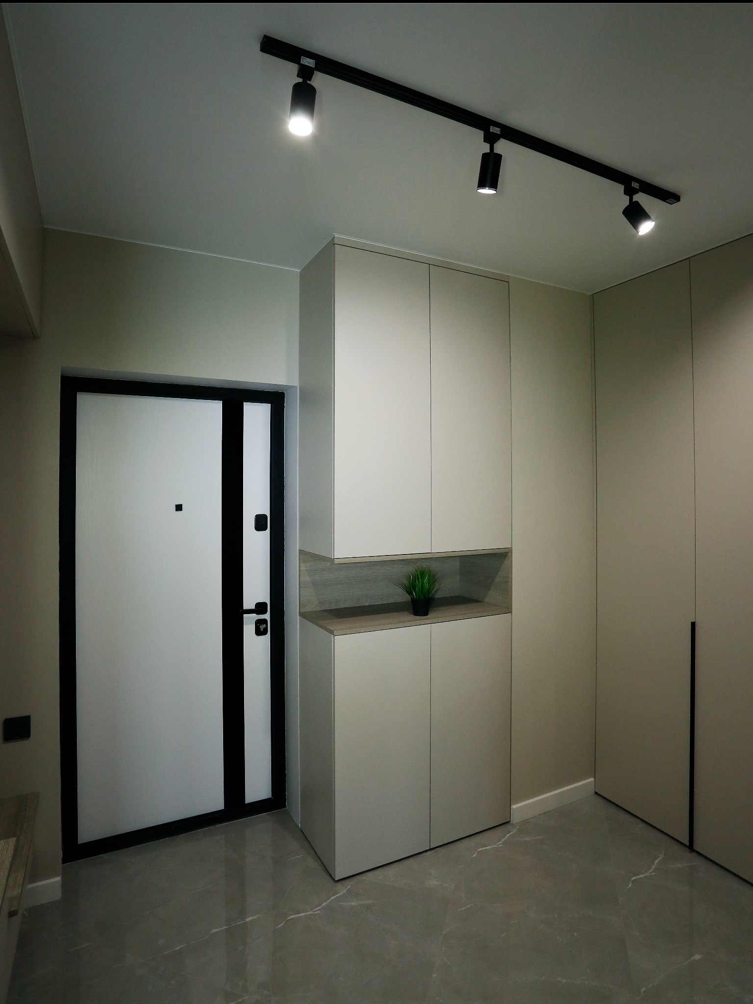 Nowoczesny przedpokój z jasnymi szafami, czarną ramą drzwi wejściowych i oświetleniem szynowym.