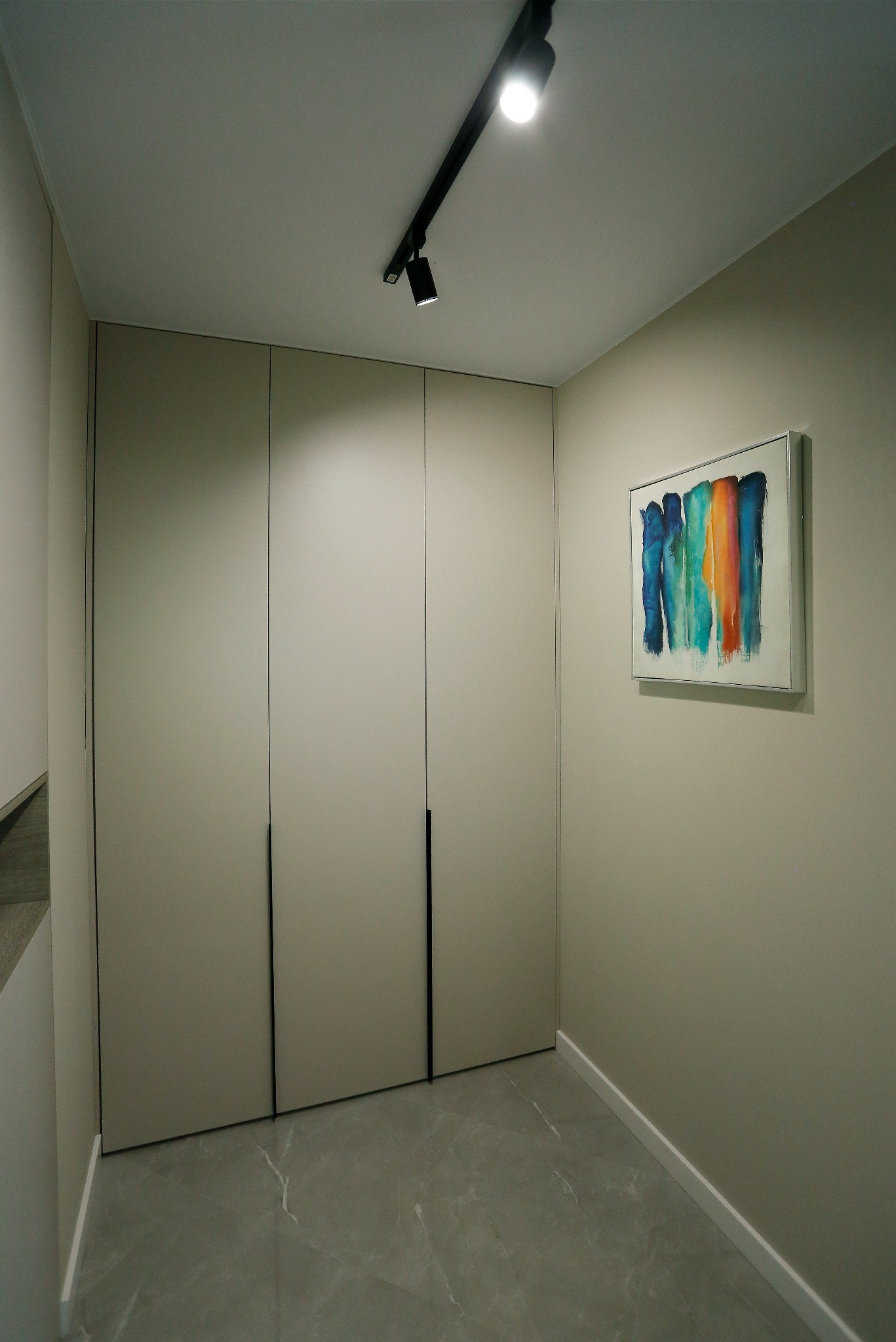 Beżowa zabudowa szafy z minimalistycznymi, czarnymi uchwytami, oświetlona reflektorem szynowym; na ścianie abstrakcyjny obraz z pionowymi smugami w odcieniach niebieskiego, zielonego i pomarańczowego.