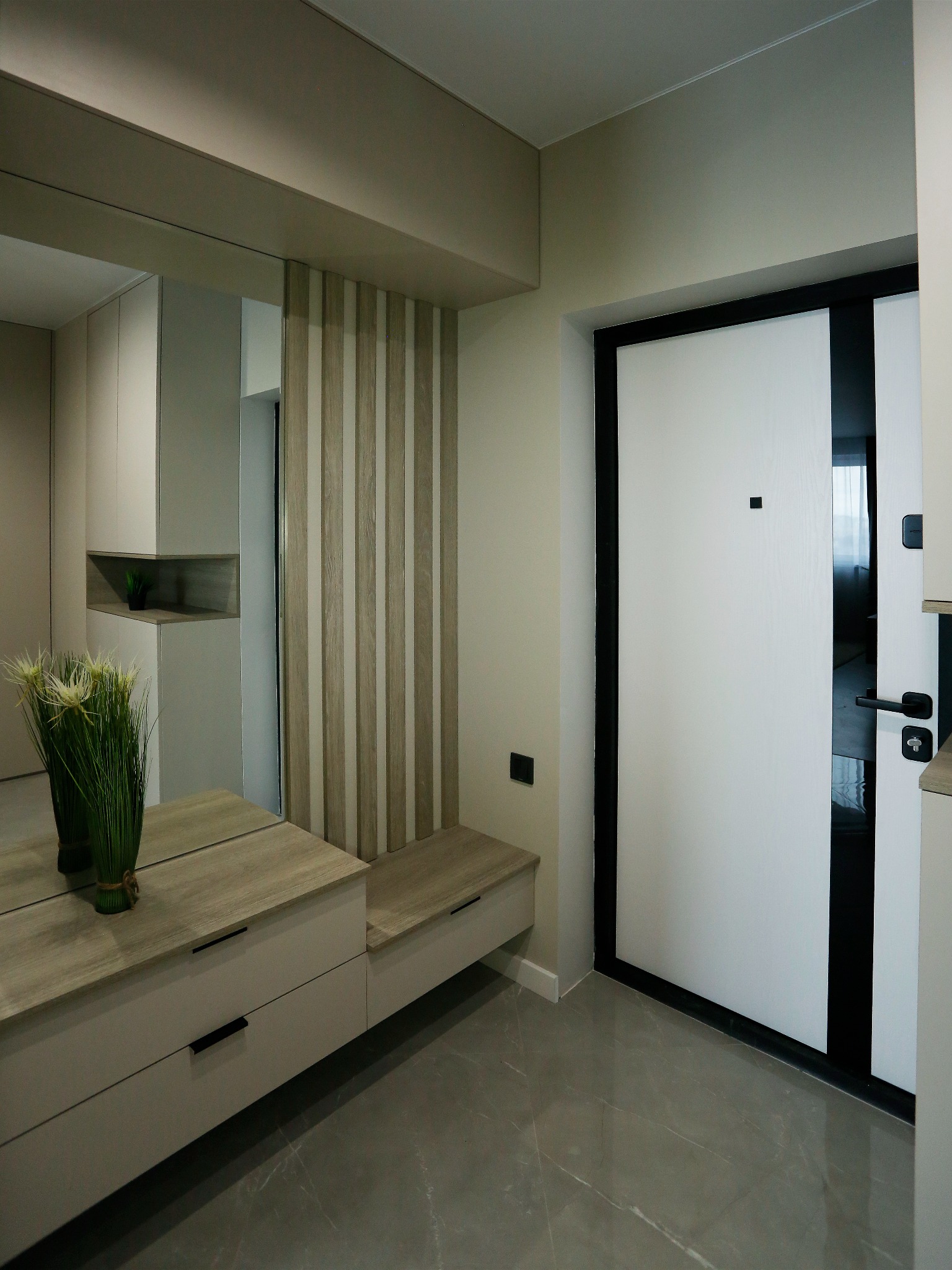 Nowoczesny przedpokój z lustrem, szafką z szufladami i siedziskiem, drzwi wejściowe w kolorze białym z czarnymi akcentami, szara marmurowa podłoga.