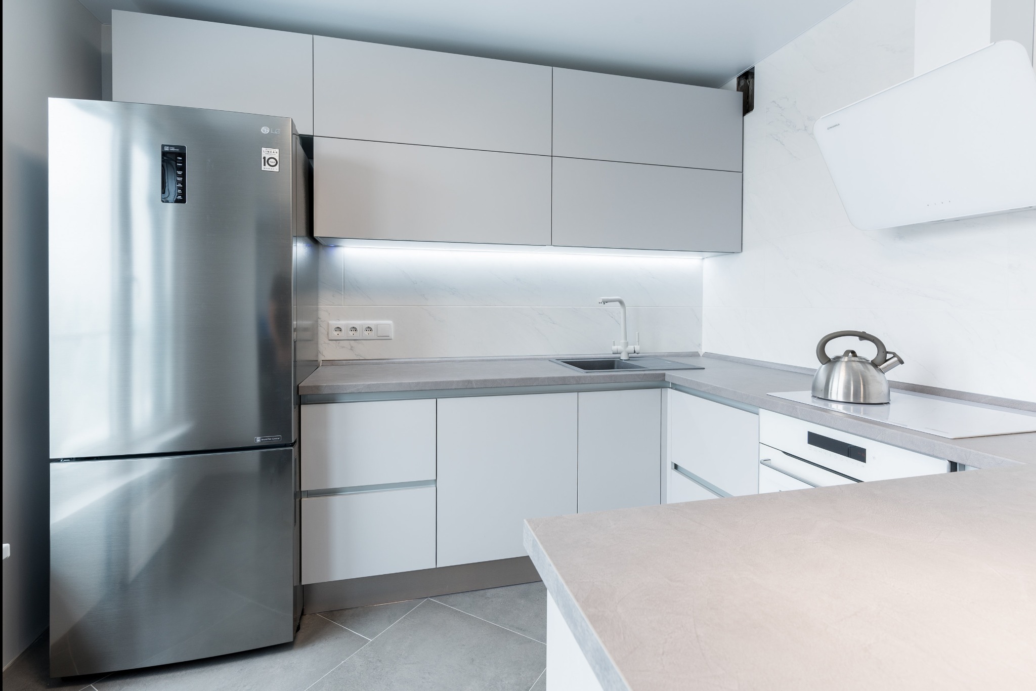 Nowoczesna, jasna kuchnia z białymi szafkami, szarym blatem, srebrną lodówką LG i czajnikiem na płycie indukcyjnej.