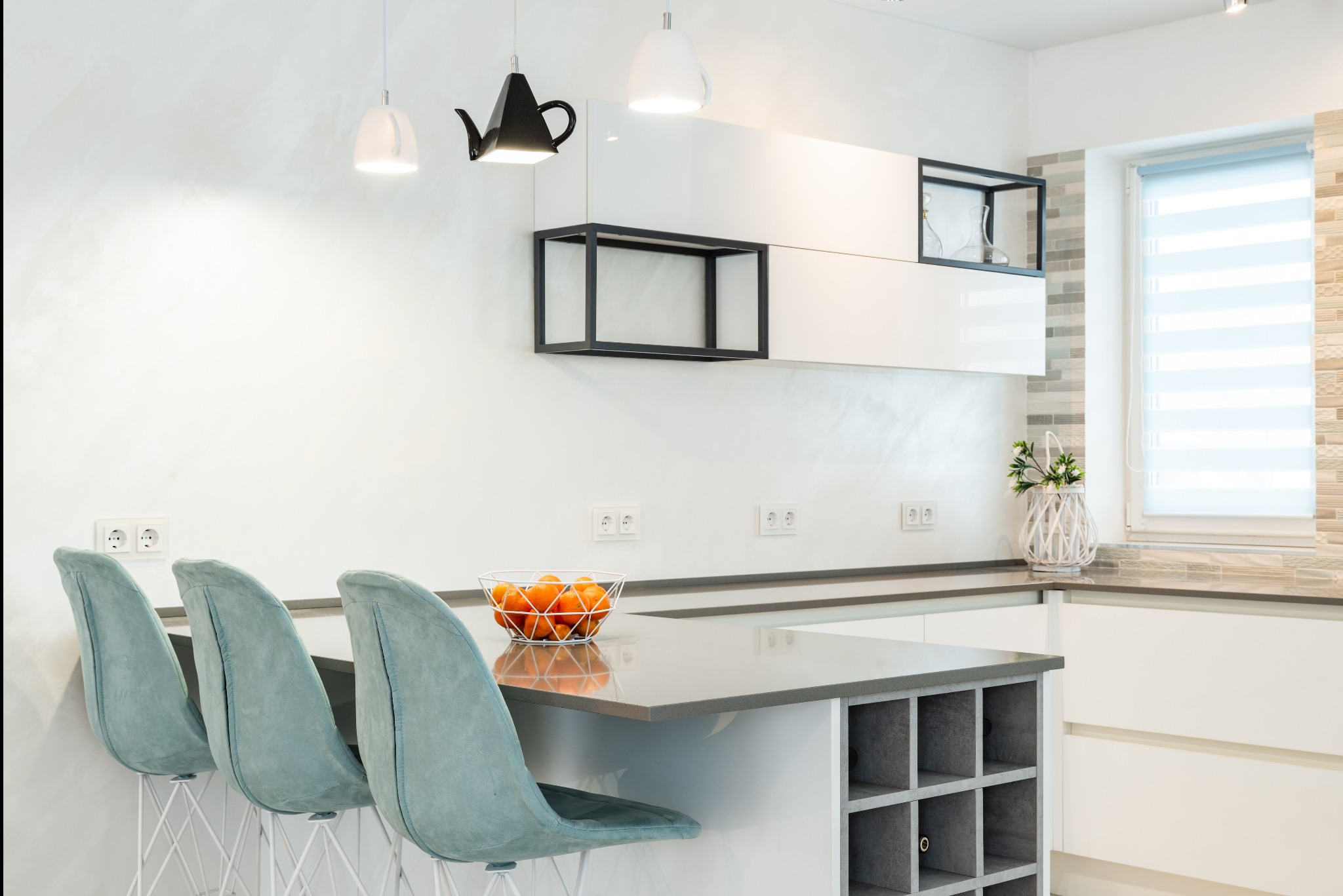 Nowoczesna, minimalistyczna kuchnia z białymi szafkami, szarym blatem, trzema turkusowymi krzesłami barowymi, designerską lampą w kształcie czajnika i misą pomarańczy.