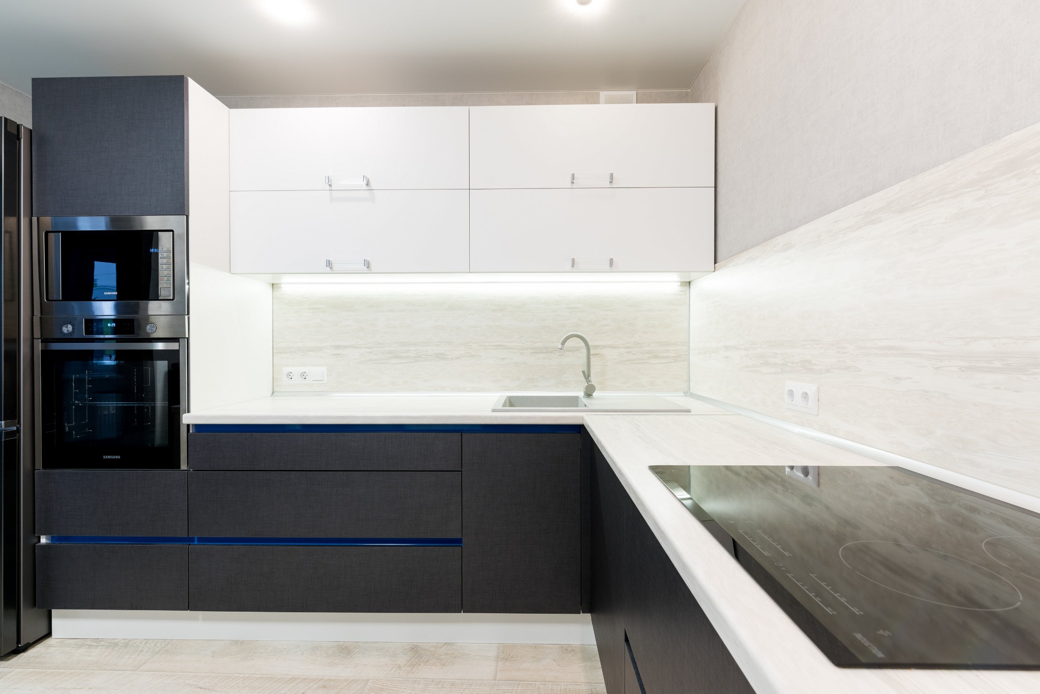 Nowoczesna kuchnia z szarymi dolnymi szafkami z niebieskimi akcentami, białymi górnymi szafkami, wbudowanym piekarnikiem i mikrofalówką Samsung oraz płytą indukcyjną.