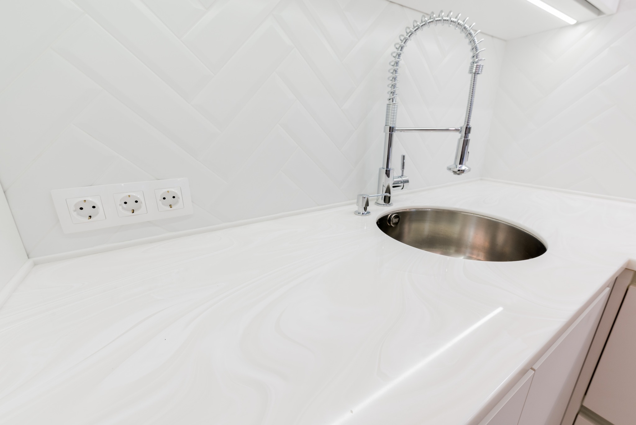 Biały blat kuchenny z marmurkowym wzorem, okrągły zlew ze stali nierdzewnej i chromowana bateria z elastyczną sprężyną, białe płytki ułożone w jodełkę na ścianie, gniazdko elektryczne z trzema...