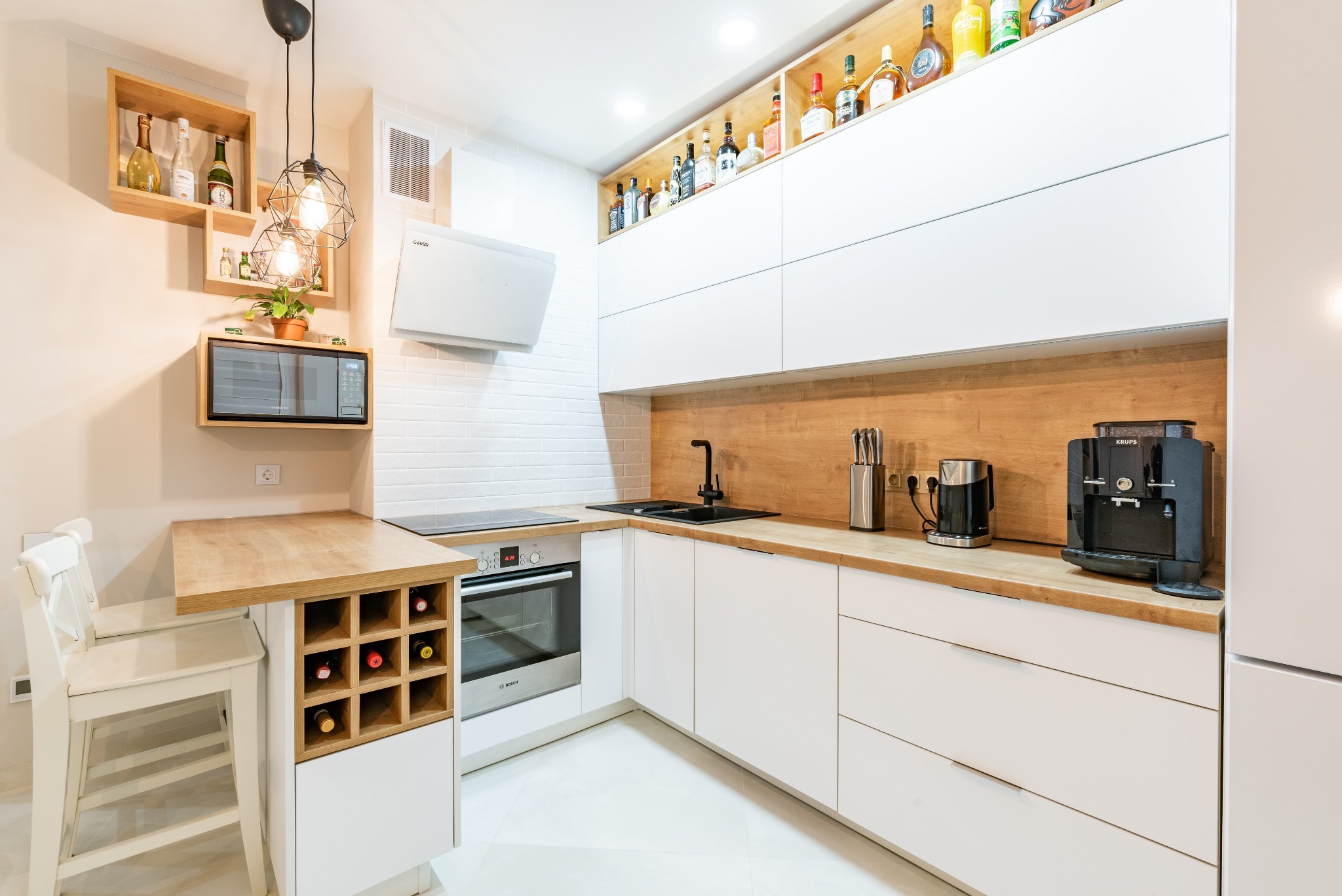 Jasna kuchnia z białymi, gładkimi frontami szafek i drewnianym blatem, z półkami na wino i alkohole, oświetlona lampami z żarówkami Edisona.