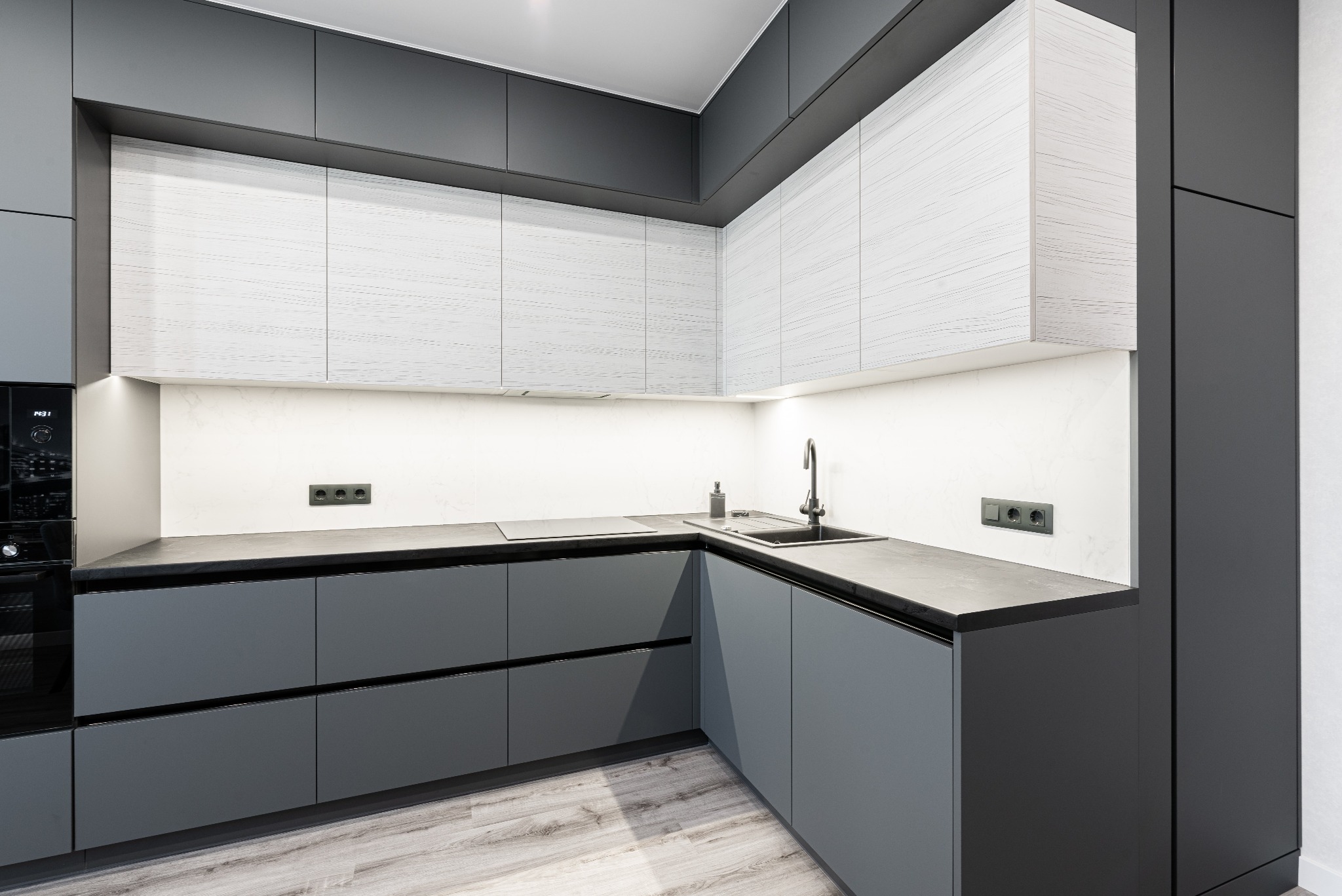 Nowoczesna, szara kuchnia z białymi szafkami górnymi w Józefowie, oświetlenie LED pod szafkami, minimalistyczny design, czarny piekarnik w zabudowie.