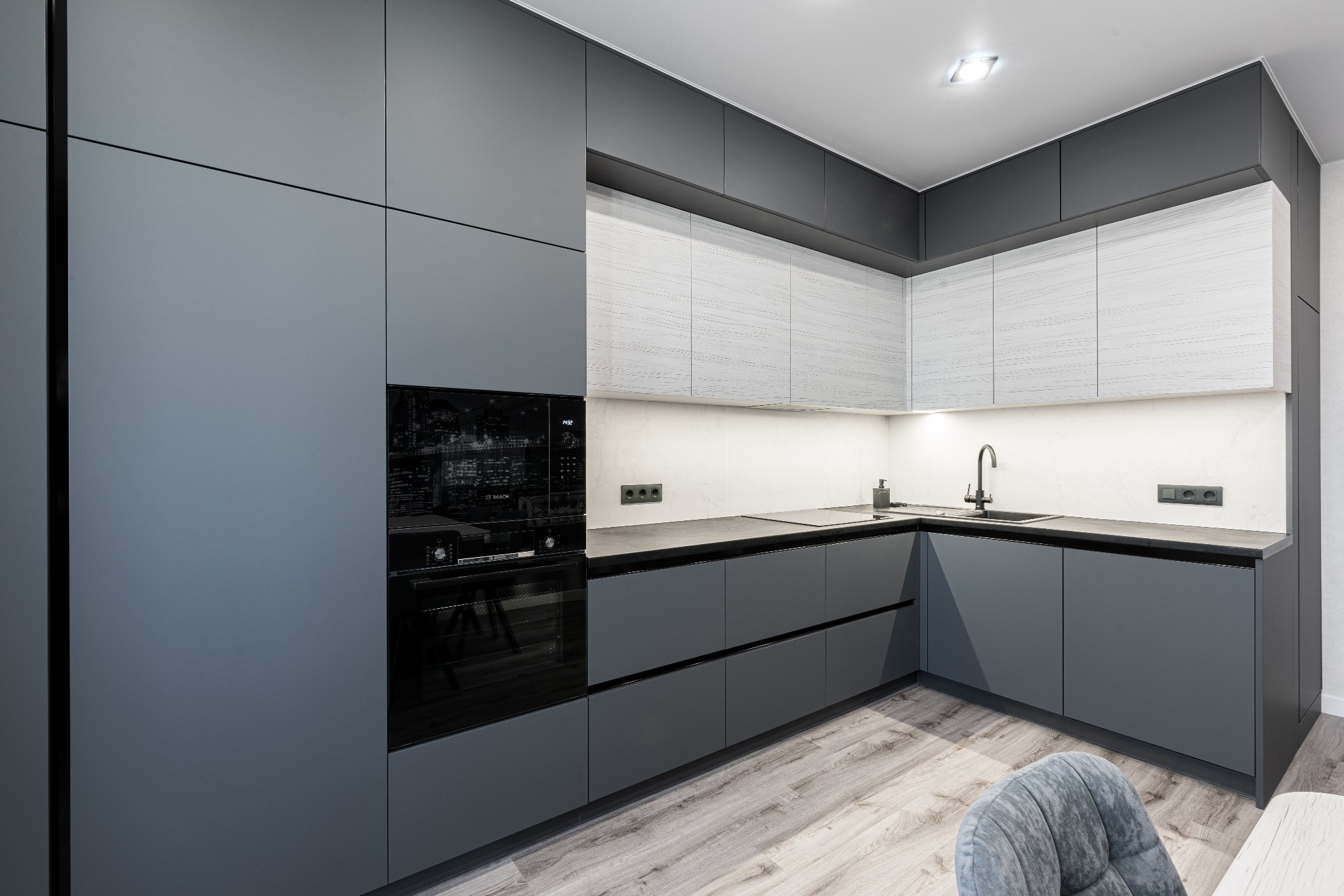 Nowoczesna, narożna kuchnia w odcieniach szarości z zabudowanym piekarnikiem Bosch, drewnopodobna podłoga i minimalistyczna armatura.