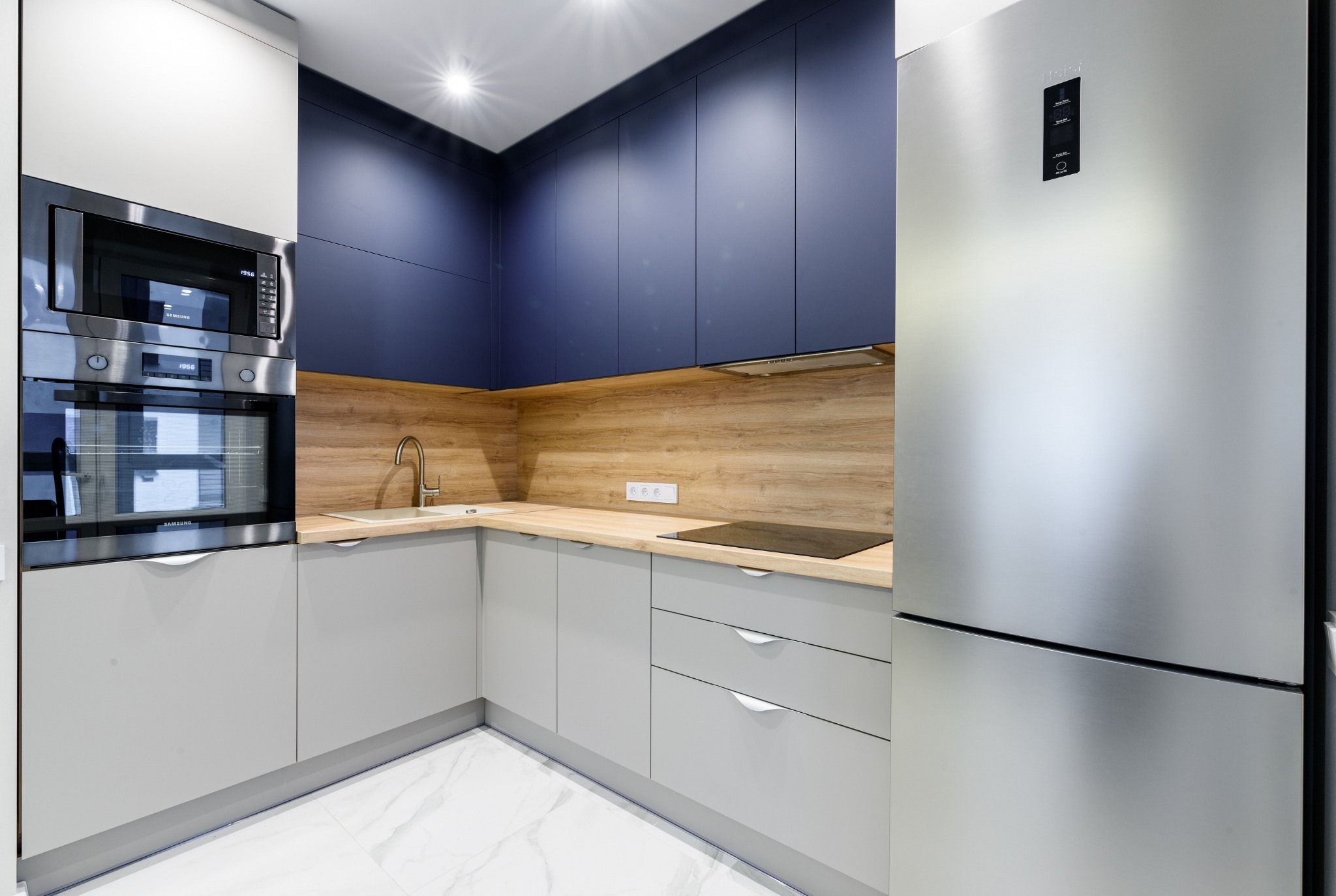 Nowoczesna kuchnia z szarymi szafkami dolnymi i ciemnoniebieskimi górnymi, drewnianym blatem i srebrną lodówką, oświetlona punktowo