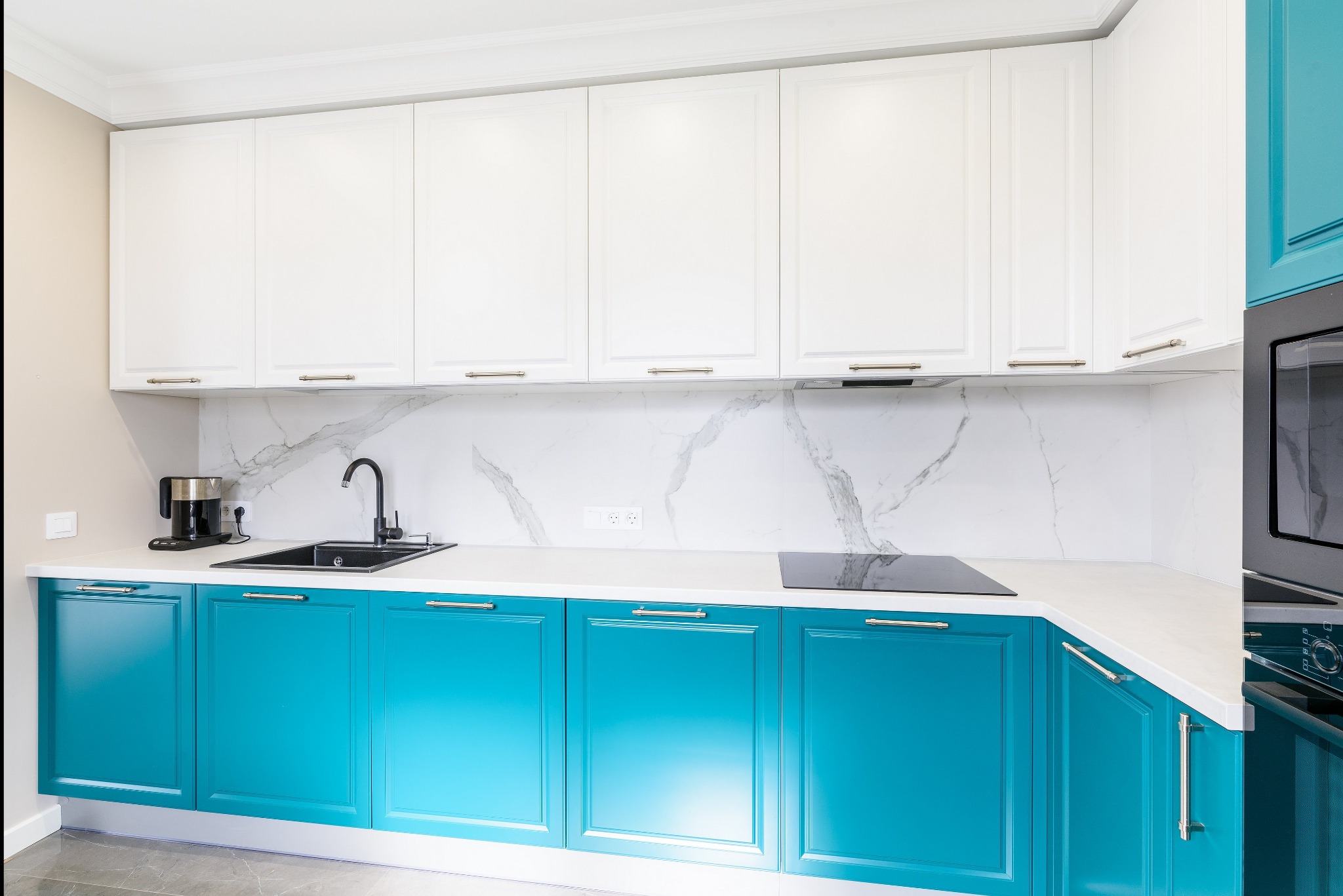 Nowoczesna kuchnia z turkusowymi szafkami dolnymi i białymi górnymi, czarnym zlewem i baterią, marmurowym backsplashiem oraz sprzętem AGD w zabudowie.