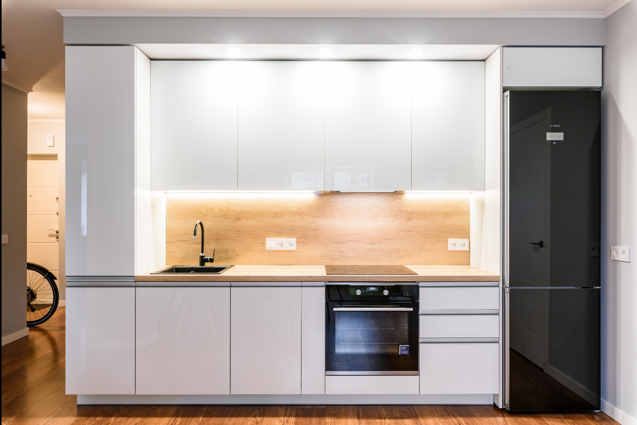 Nowoczesna, biała zabudowa kuchenna z drewnianym blatem i czarnym zlewem, oświetlenie LED pod szafkami wiszącymi, czarna lodówka z wyświetlaczem, widok na fragment roweru w tle.