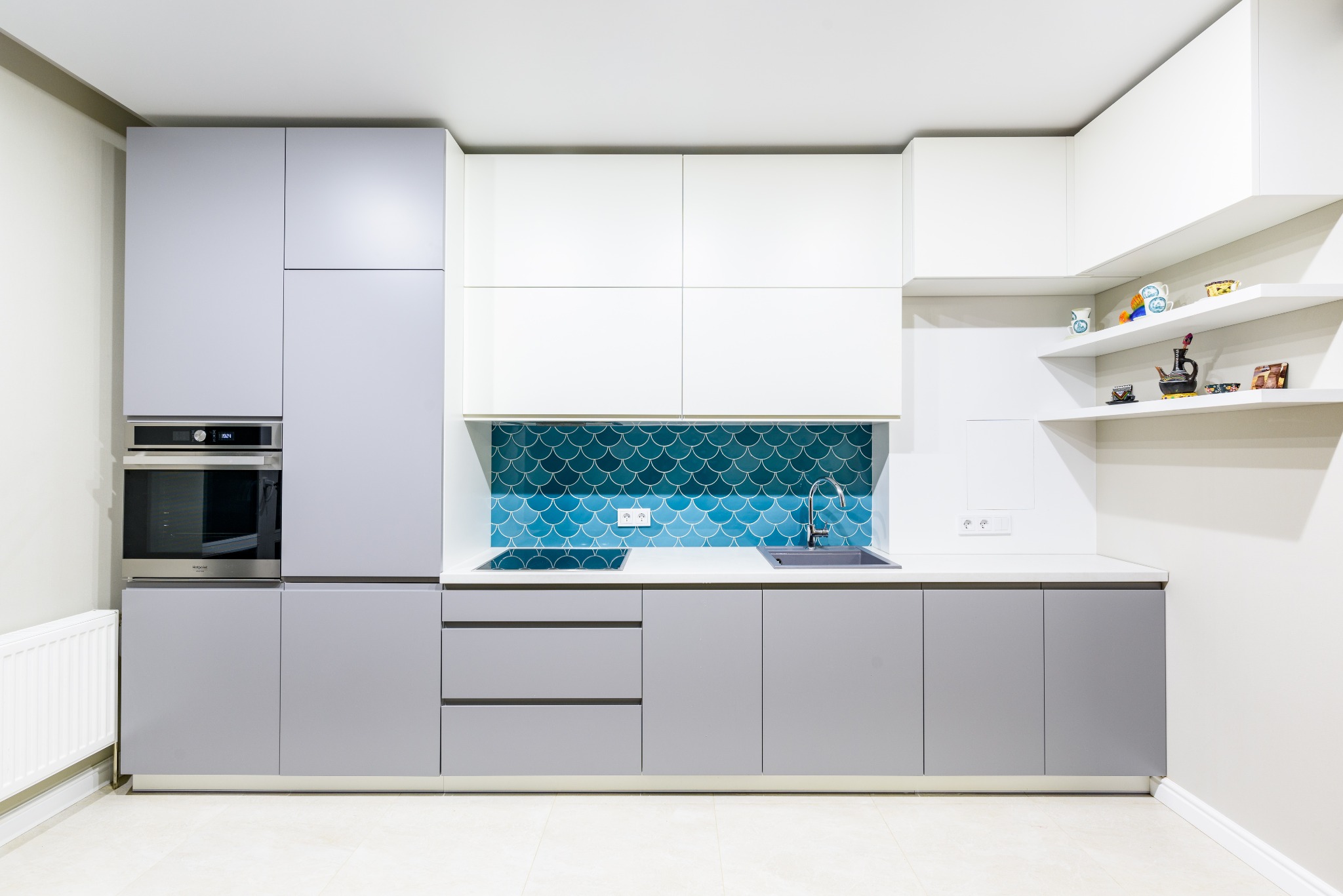 Nowoczesna kuchnia z szarymi i białymi szafkami, turkusowymi płytkami w kształcie rybiej łuski nad blatem, piekarnikiem w zabudowie i otwartymi półkami z dekoracjami.
