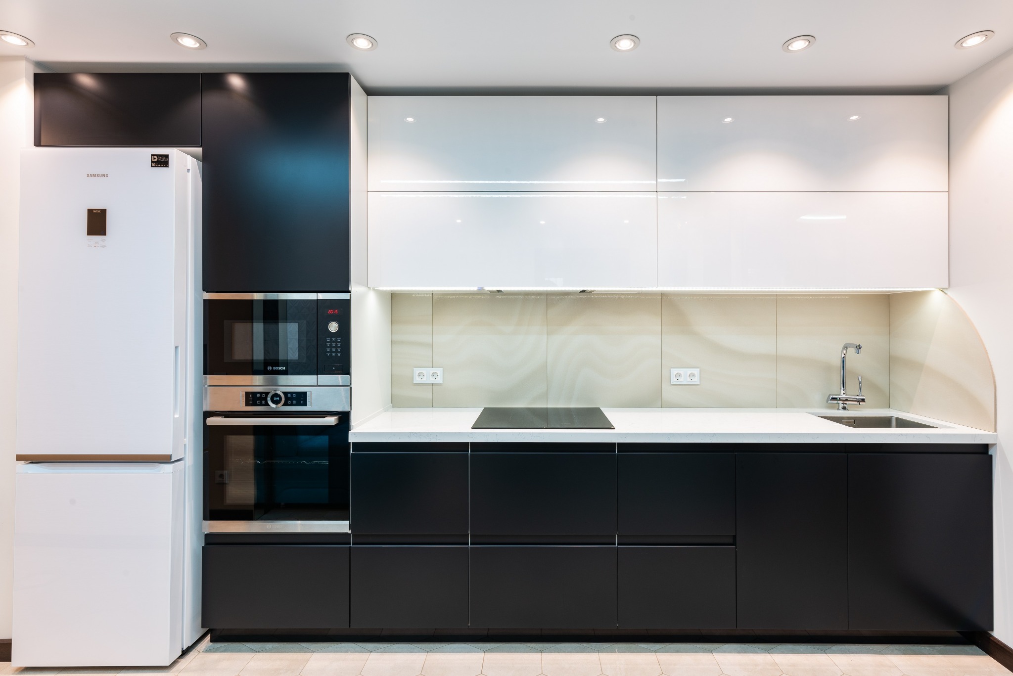 Nowoczesna kuchnia z czarnymi dolnymi szafkami i białymi górnymi, biała lodówka Samsung i piekarnik Bosch w zabudowie, jasny blat i płytki na ścianie