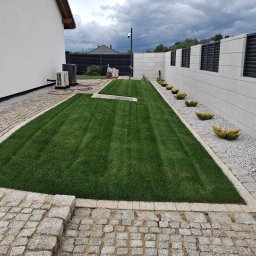 M-Garden Usługi Ogrodnicze Marcin Łacny - Zadbany trawnik z rolki, obrzeża z kostki granitowej, ozdobne krzewy wzdłuż betonowego ogrodzenia. Widok na nowoczesny dom i pochmurne niebo.