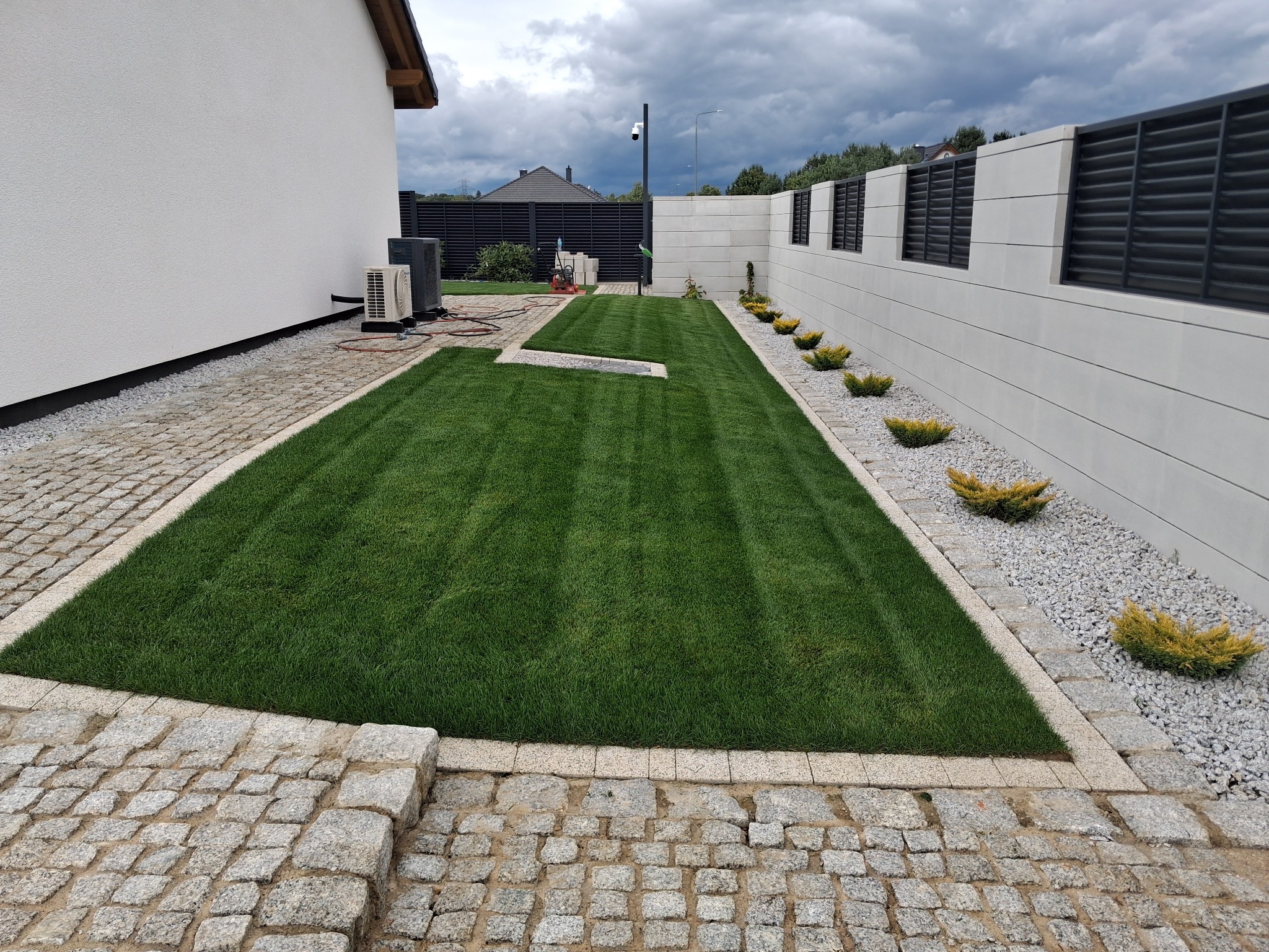 Zadbany trawnik z rolki, obrzeża z kostki granitowej, ozdobne krzewy wzdłuż betonowego ogrodzenia. Widok na nowoczesny dom i pochmurne niebo.