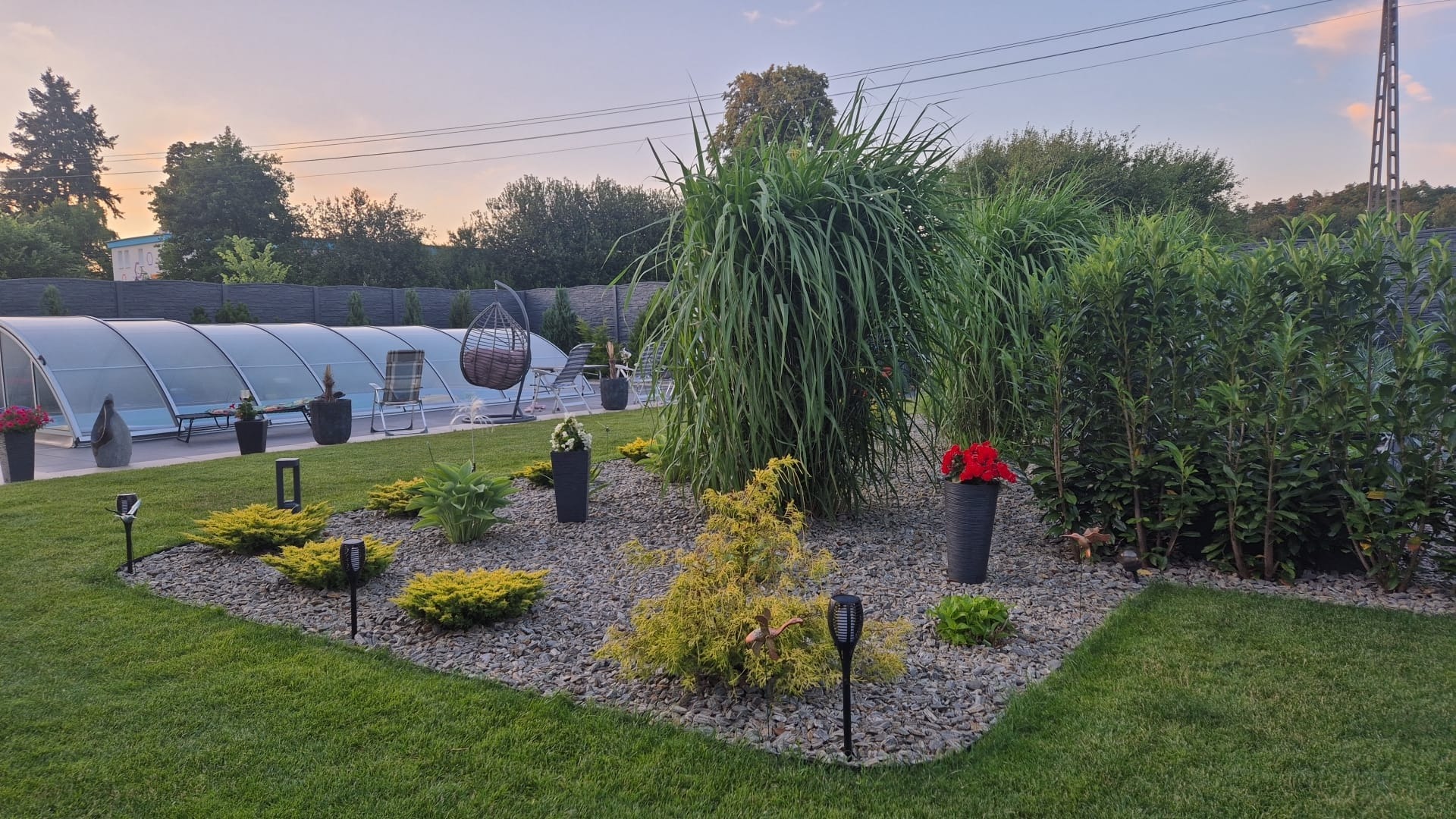 Urozmaicony ogród z ozdobnymi trawami, krzewami i kwiatami w donicach. W tle zadaszony basen i wiszące krzesło. Oświetlenie punktowe podkreśla aranżację.