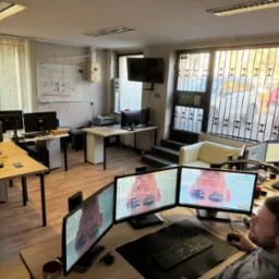 Wnętrze biura IT z kilkoma stanowiskami pracy, gdzie programista pracuje na trzech monitorach, wyświetlających zdjęcie osoby w okularach przeciwsłonecznych, a na ścianie wisi tablica z rysunkami...