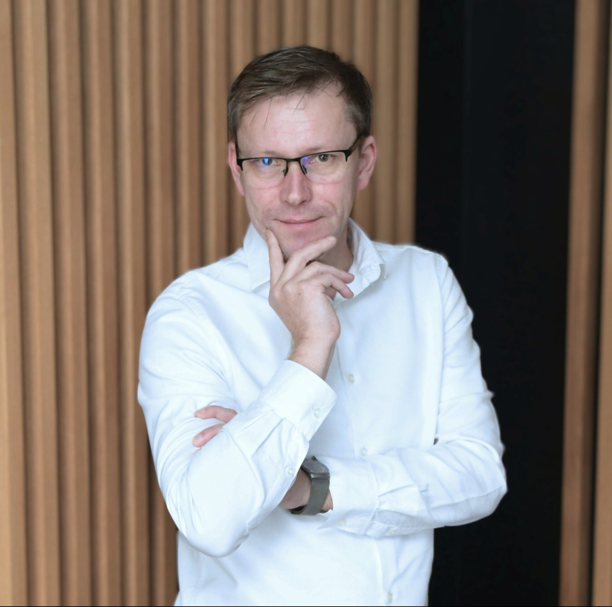 Portret mężczyzny w okularach, w białej koszuli, z ręką podpartą pod brodą, na tle drewnianej ściany z czarnym elementem.