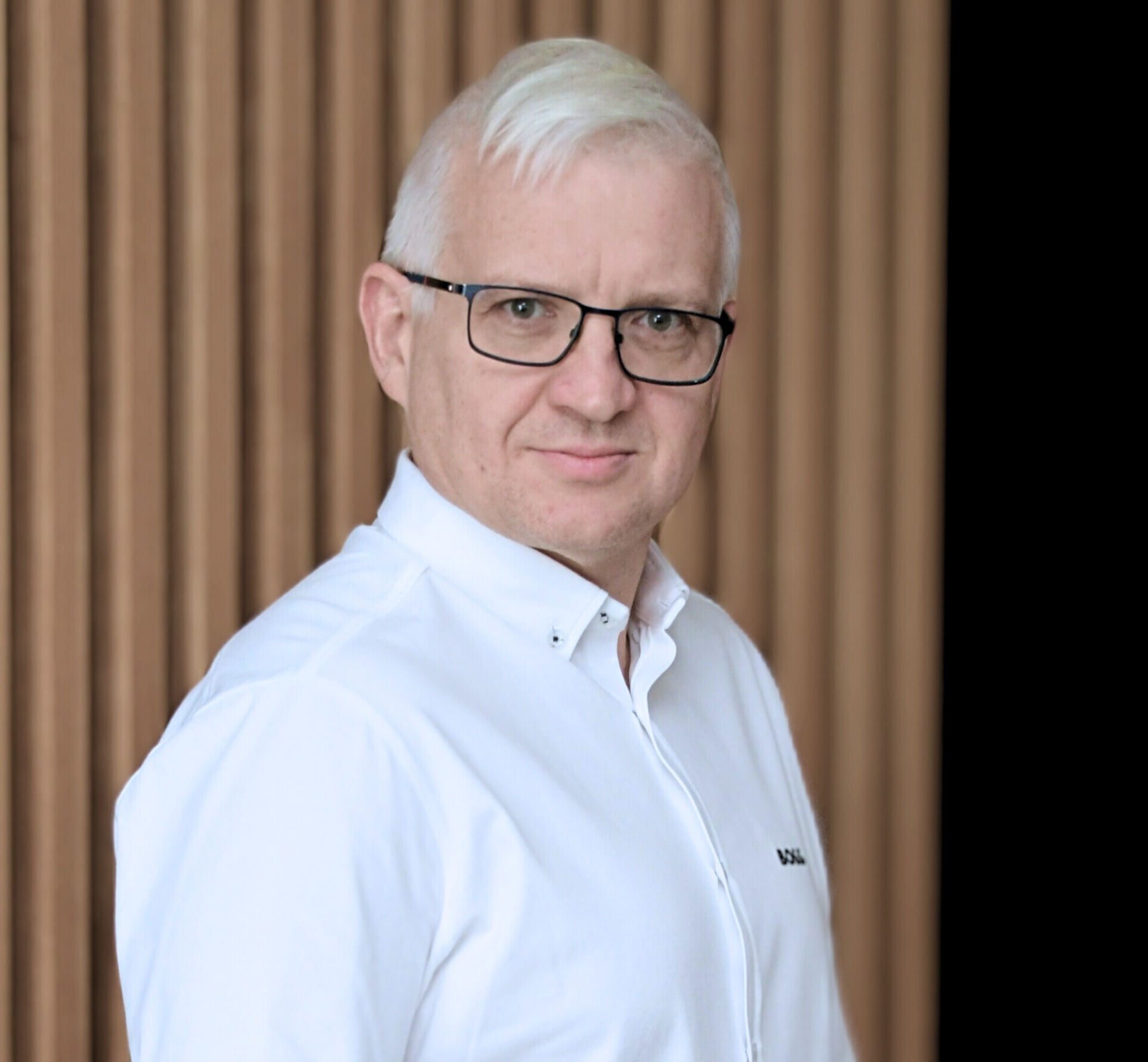 Portret mężczyzny w okularach i białej koszuli na tle drewnianej ściany, uśmiechający się lekko.