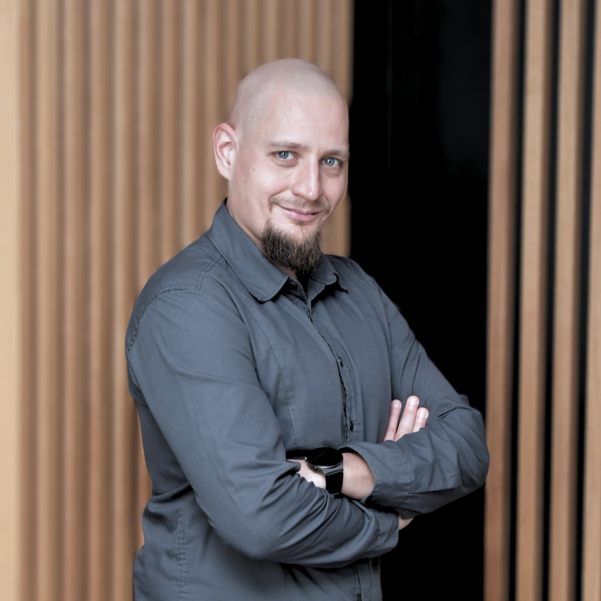 Portret uśmiechniętego mężczyzny z brodą i zegarkiem na tle drewnianej ściany, sugerujący profesjonalizm w branży IT.