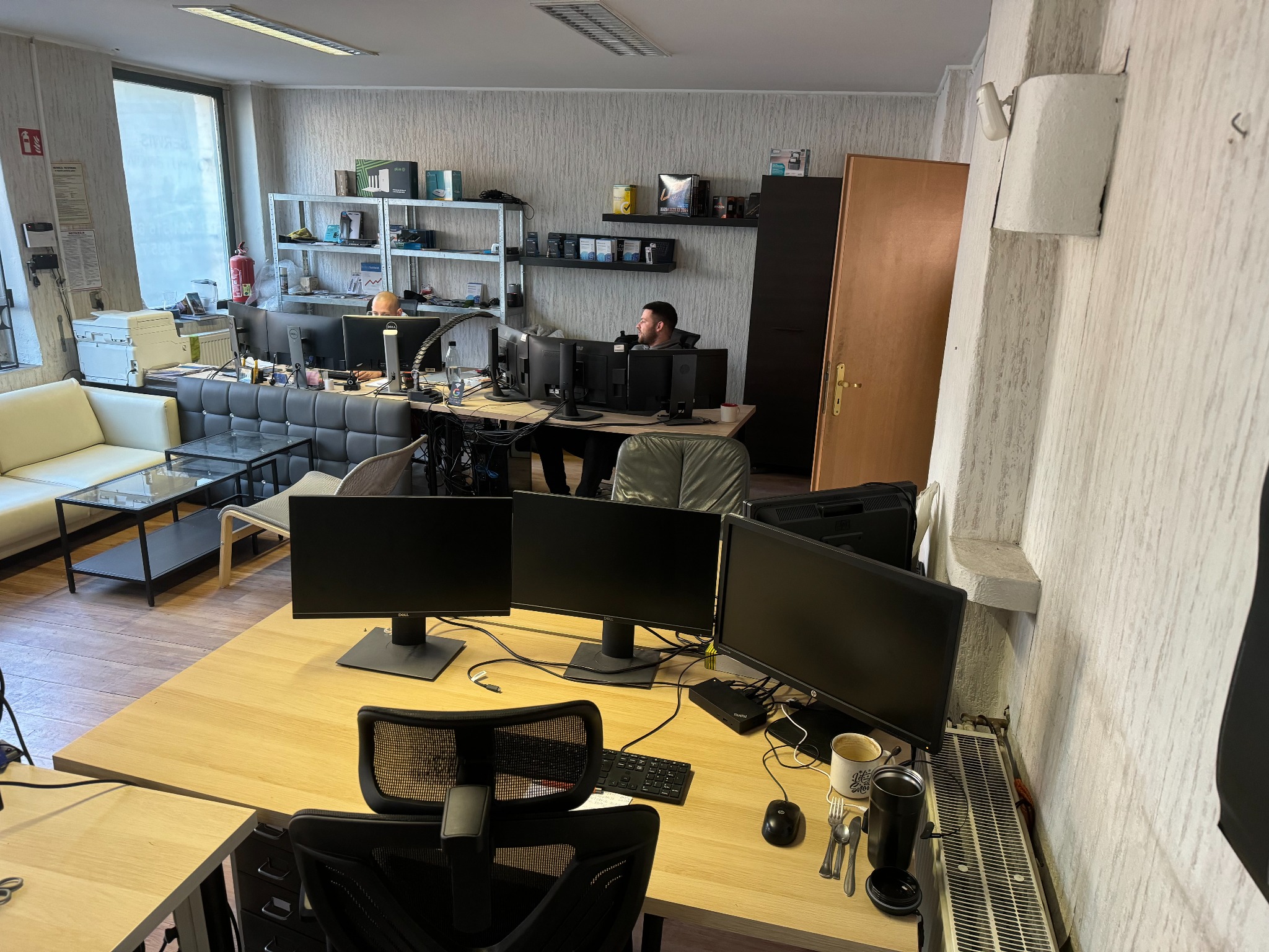 Biuro z wieloma stanowiskami komputerowymi, pracownik siedzi przy jednym z nich, otoczony kilkoma monitorami, na biurku widoczne akcesoria i kubek, w tle regały z urządzeniami IT.