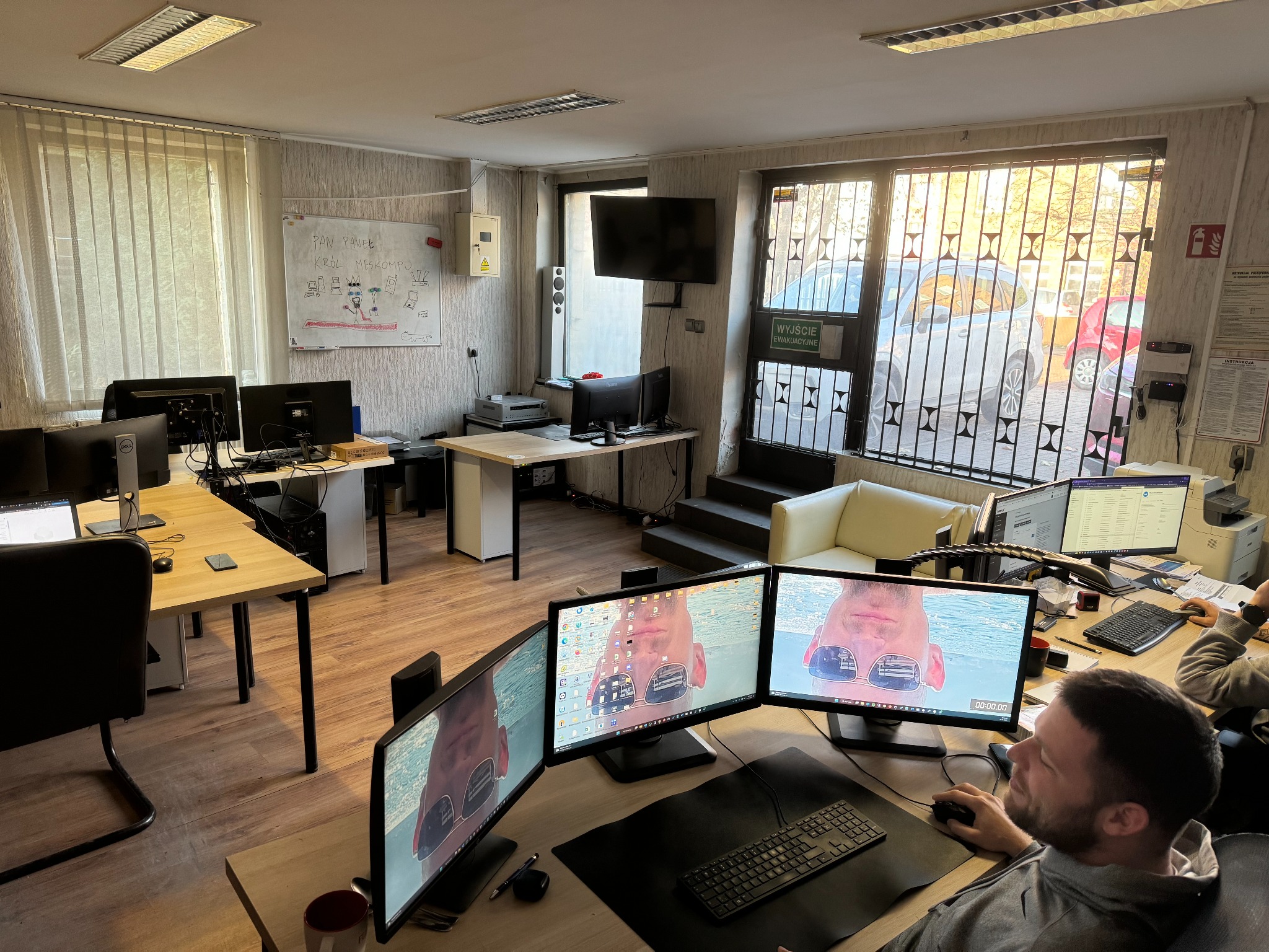 Wnętrze biura IT z kilkoma stanowiskami pracy, gdzie programista pracuje na trzech monitorach, wyświetlających zdjęcie osoby w okularach przeciwsłonecznych, a na ścianie wisi tablica z rysunkami...