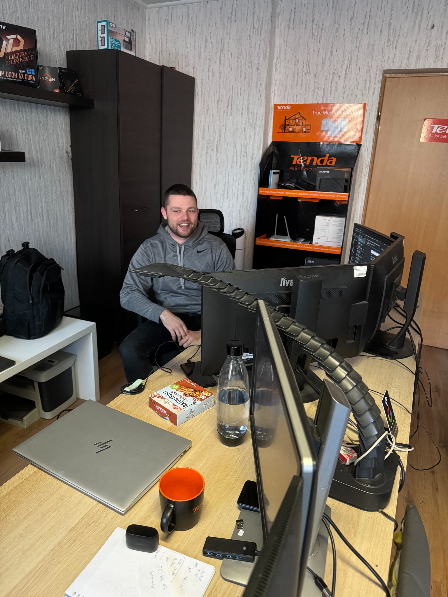 Uśmiechnięty mężczyzna siedzi przy biurku z trzema monitorami, laptopem HP, przekąską Baton Musli, kubkiem i butelką wody, w tle szafa i regał z routerami Tenda.