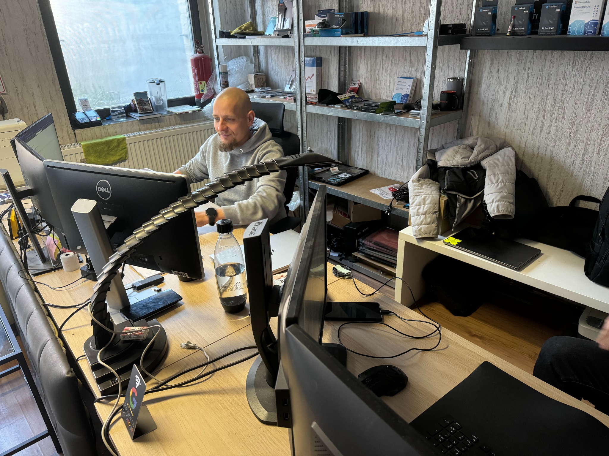 Mężczyzna z brodą i zegarkiem na nadgarstku siedzi przy biurku z trzema monitorami, butelką wody gazowanej i kartą z prośbą o recenzję w Google, w tle półki z akcesoriami komputerowymi i pudełkami...